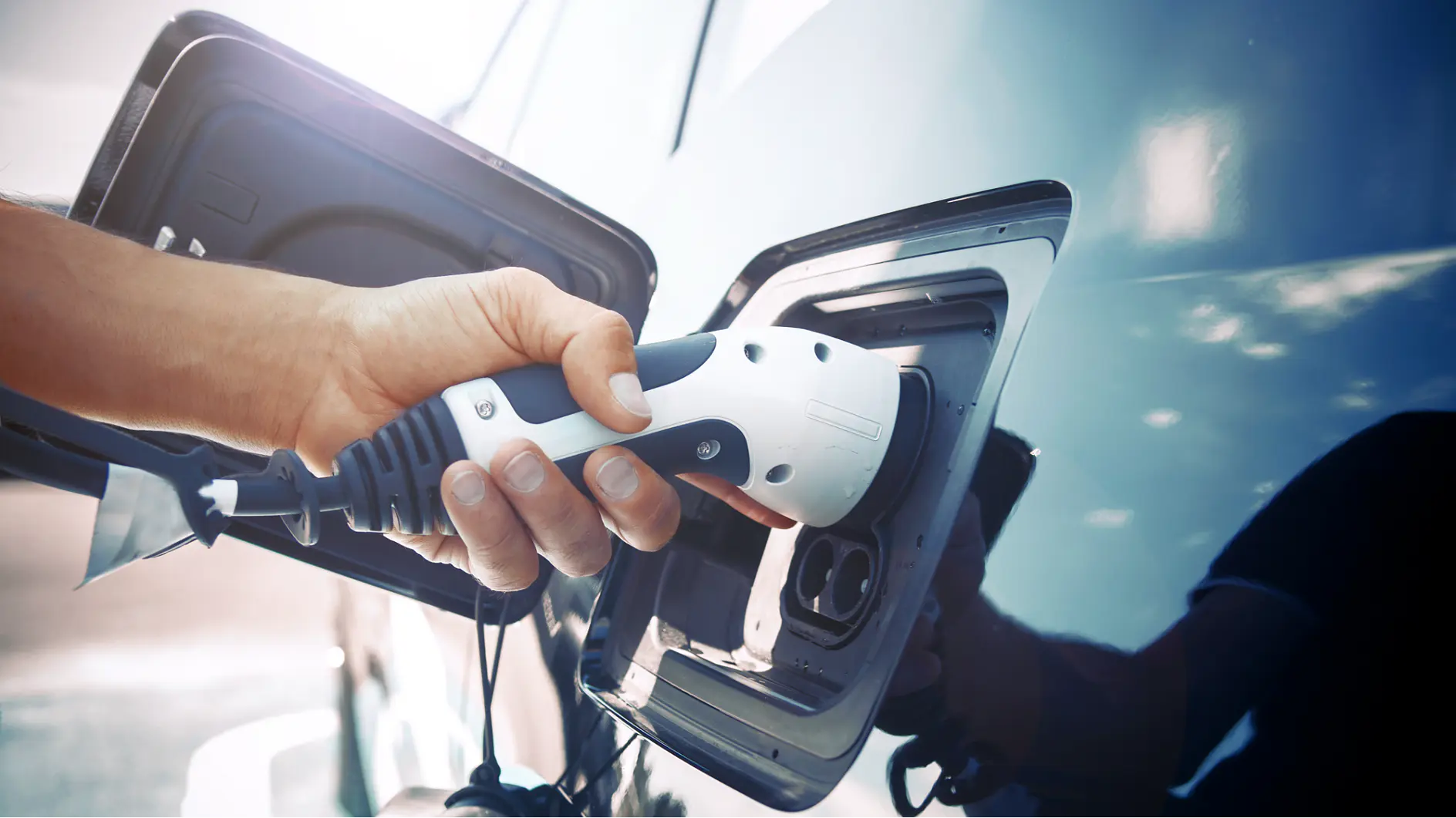 Une main branche un connecteur de recharge blanc pour véhicule électrique dans le port de charge d’une voiture bleue. Le port de charge de la voiture est ouvert et la lumière du soleil se reflète sur la surface du véhicule, avec un rouleau de ruban tesa à proximité pour tout besoin de fixation.