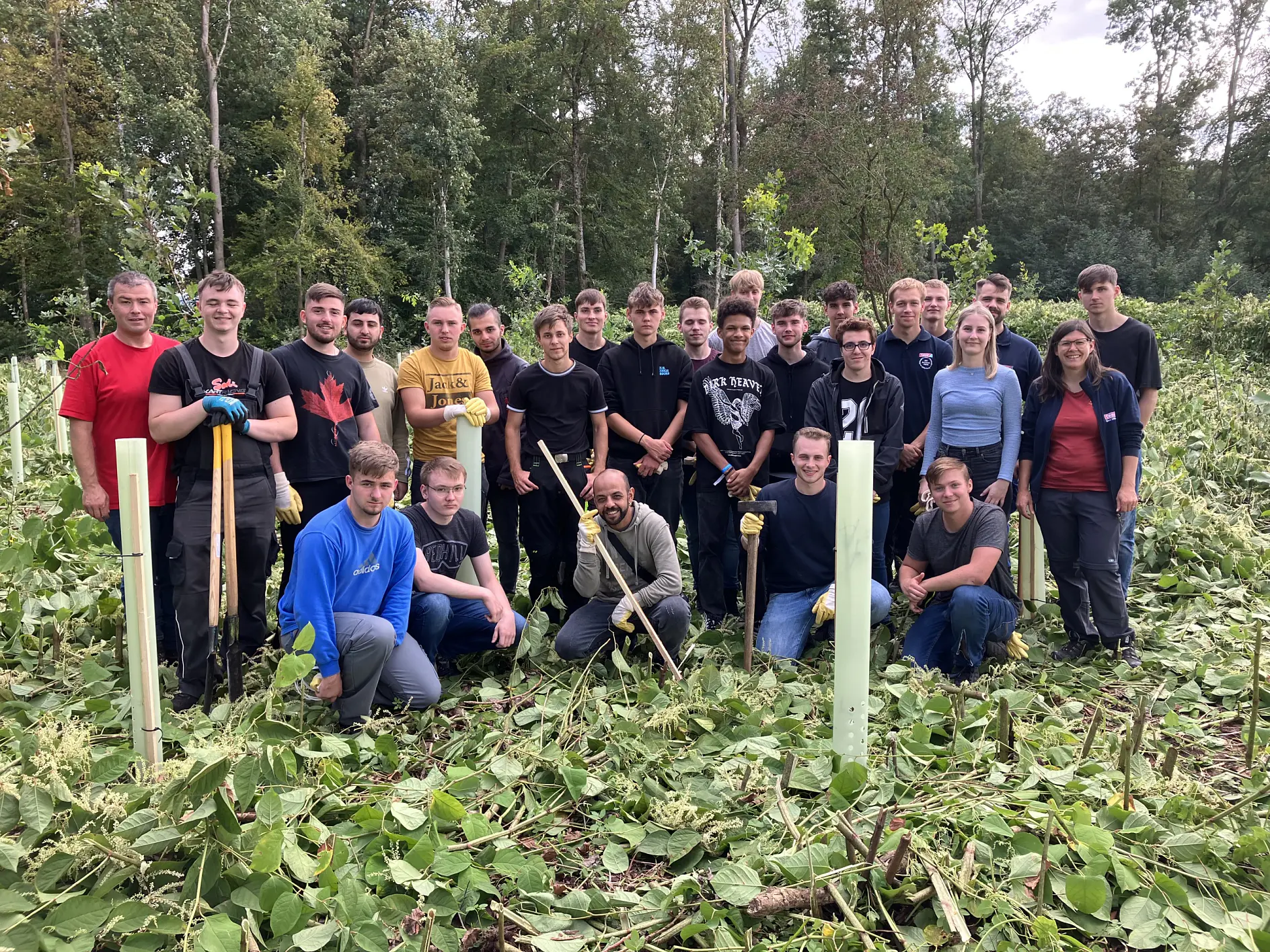 Un groupe de personnes est rassemblé dans une zone boisée avec de la végétation verte. Ils sont habillés de manière décontractée, certains tenant du ruban tesa et des outils. À l'arrière-plan, on voit des arbres et un ciel nuageux.