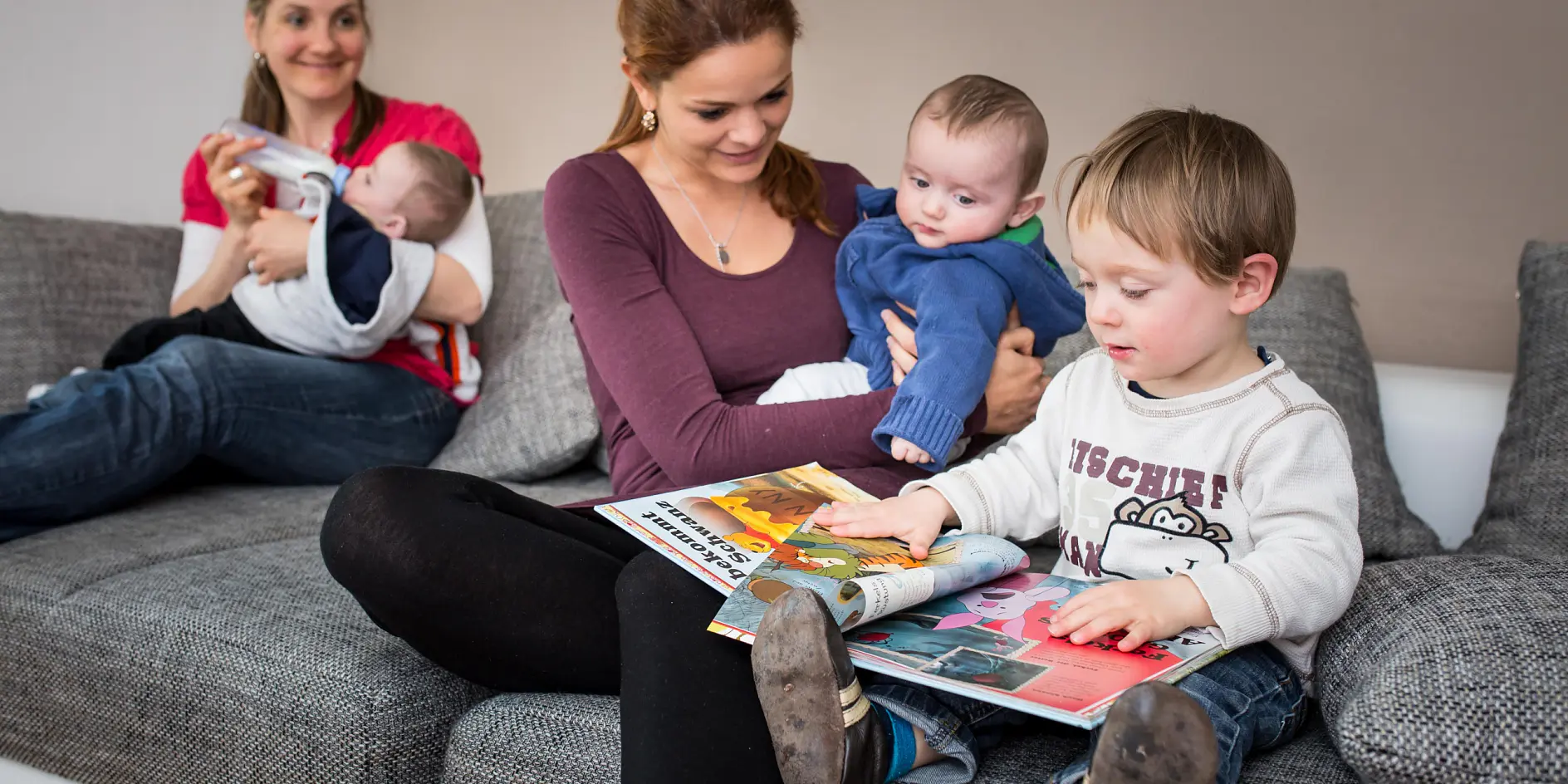 Une femme tient un bébé tout en étant assise sur un canapé gris. Une autre femme est assise à côté d'elle et tient également un bébé. À côté d'elles, un enfant est assis, regardant un livre ouvert, près d'un rouleau de ruban tesa. L'endroit semble être un salon.
