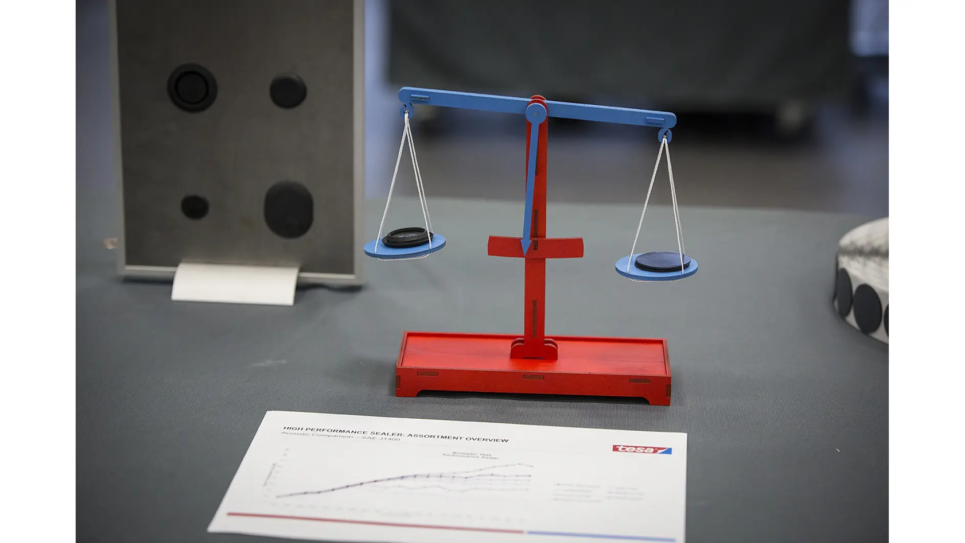 Une balance rouge et bleue tient un objet noir de chaque côté, montrant qu'ils ont le même poids. Au premier plan, une feuille avec un graphique et un logo est posée sur une surface grise, mettant en avant du ruban tesa. Les objets en arrière-plan sont flous. (This text has been generated by AI)