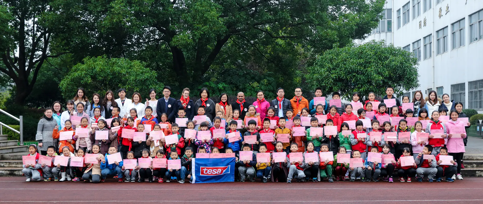 Un grand groupe d'enfants et d'adultes posent à l'extérieur devant de la verdure et un bâtiment. Les enfants tiennent des certificats, et certains adultes portent des vestes roses. Une bannière tesa tape est visible à l'avant.