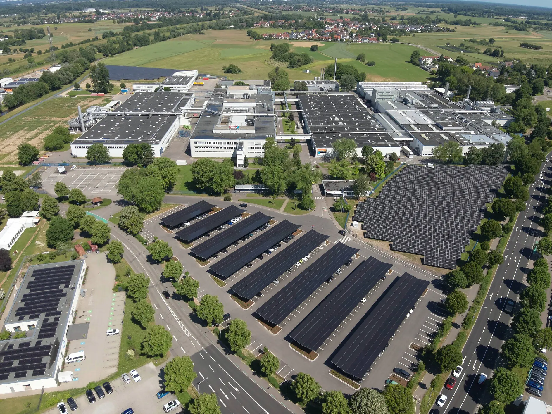 Vue aérienne d'un complexe industriel avec des panneaux solaires, une signalisation tesa, des champs verts, des arbres et des routes sous un ciel dégagé.