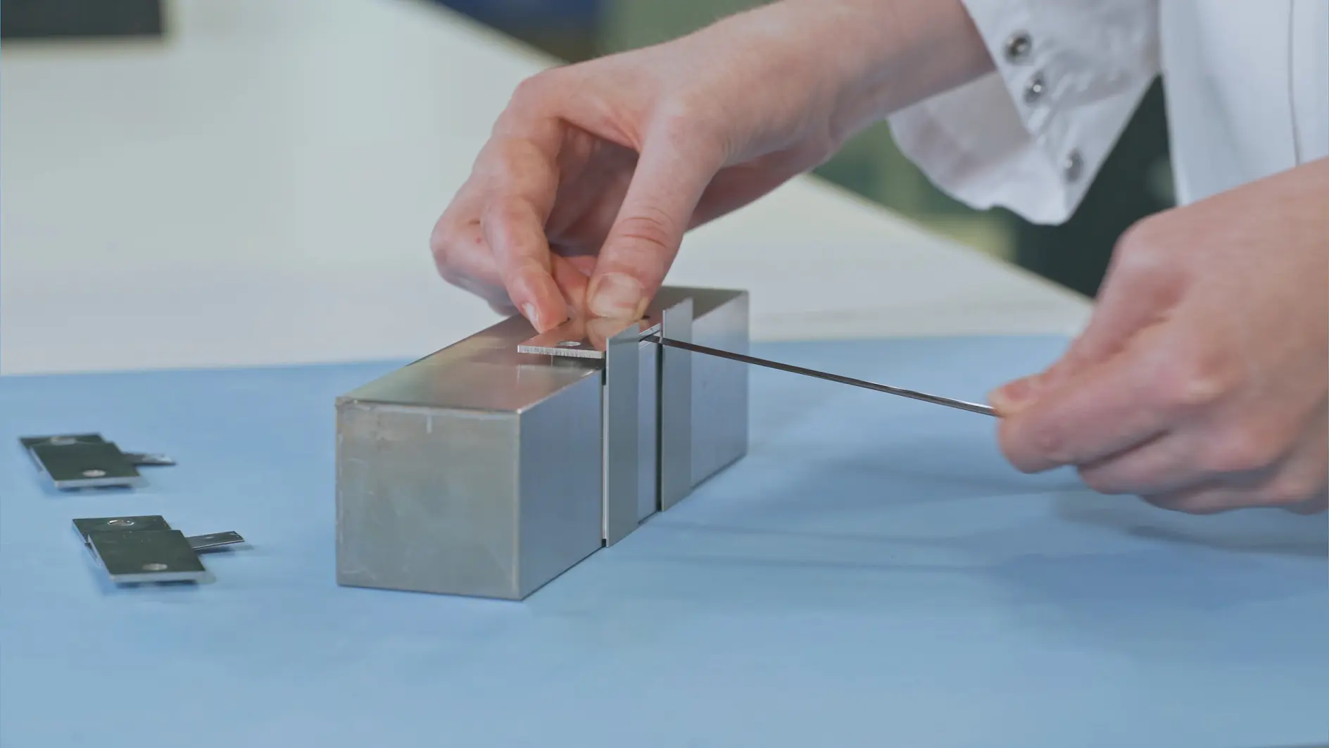 Une personne en blouse blanche utilise un fin outil métallique sur un appareil métallique rectangulaire posé sur une surface bleue. Deux pièces métalliques supplémentaires sont posées à proximité, fixées avec du ruban tesa. La scène semble se dérouler dans un laboratoire.