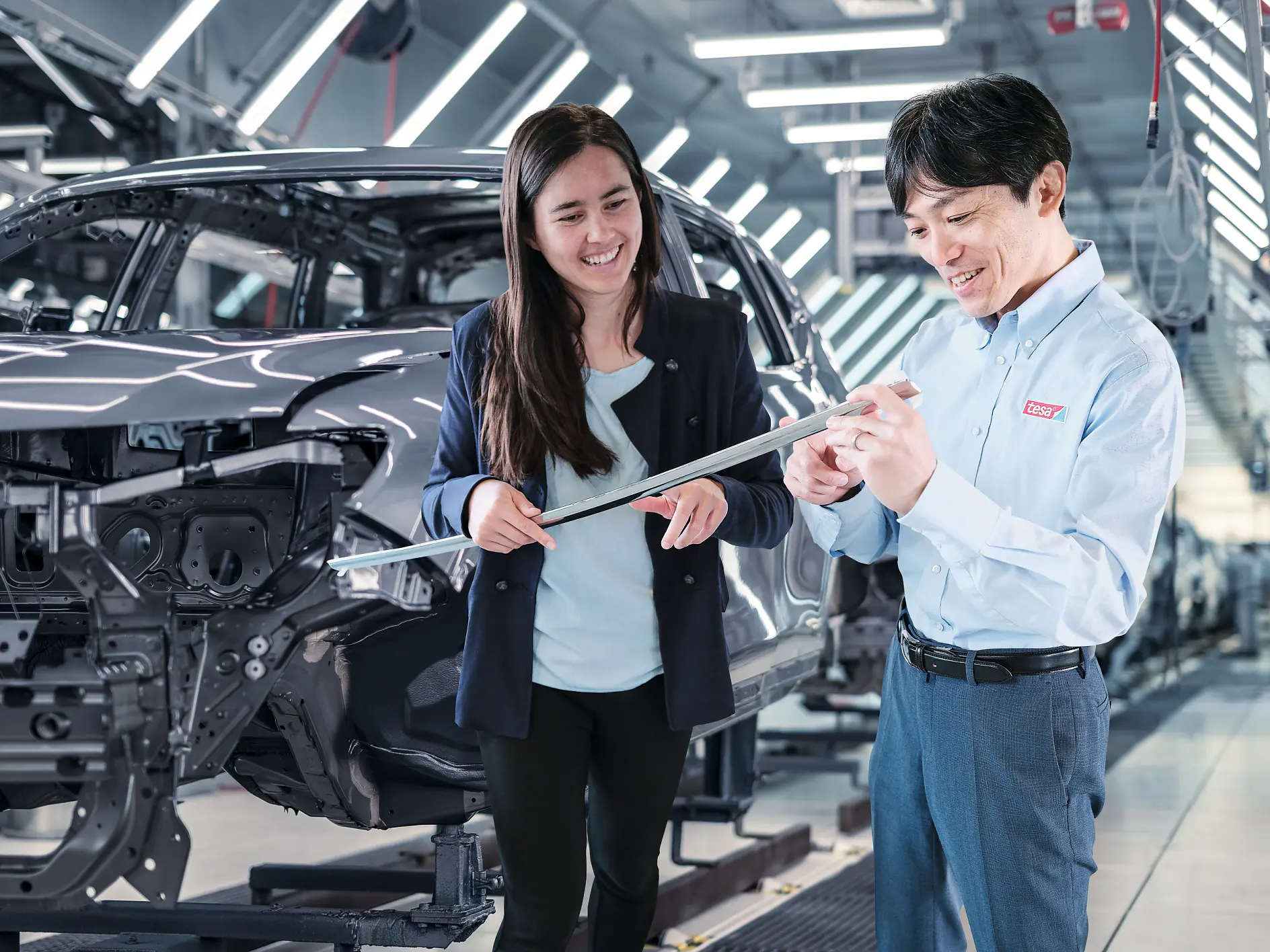 Deux personnes dans une usine automobile, près d’un véhicule partiellement assemblé, discutent de plans, avec du ruban tesa visible. Des lumières vives remplissent l’espace industriel.