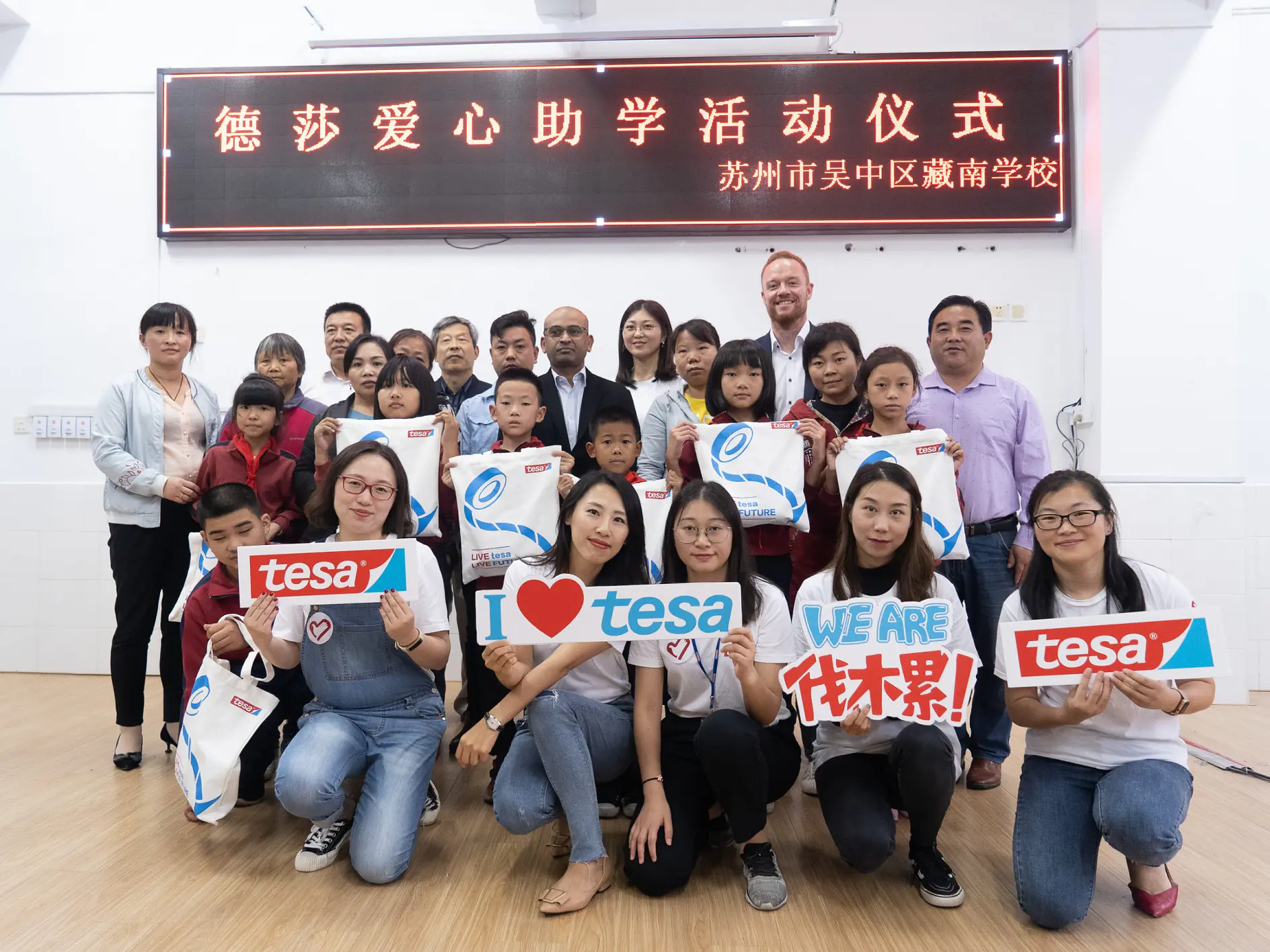 Un groupe diversifié d’adultes et d’enfants pose pour une photo de groupe à l’intérieur. Plusieurs personnes tiennent des panneaux et des sacs de marque tesa. Des caractères chinois sont affichés sur une pancarte au-dessus d’eux.