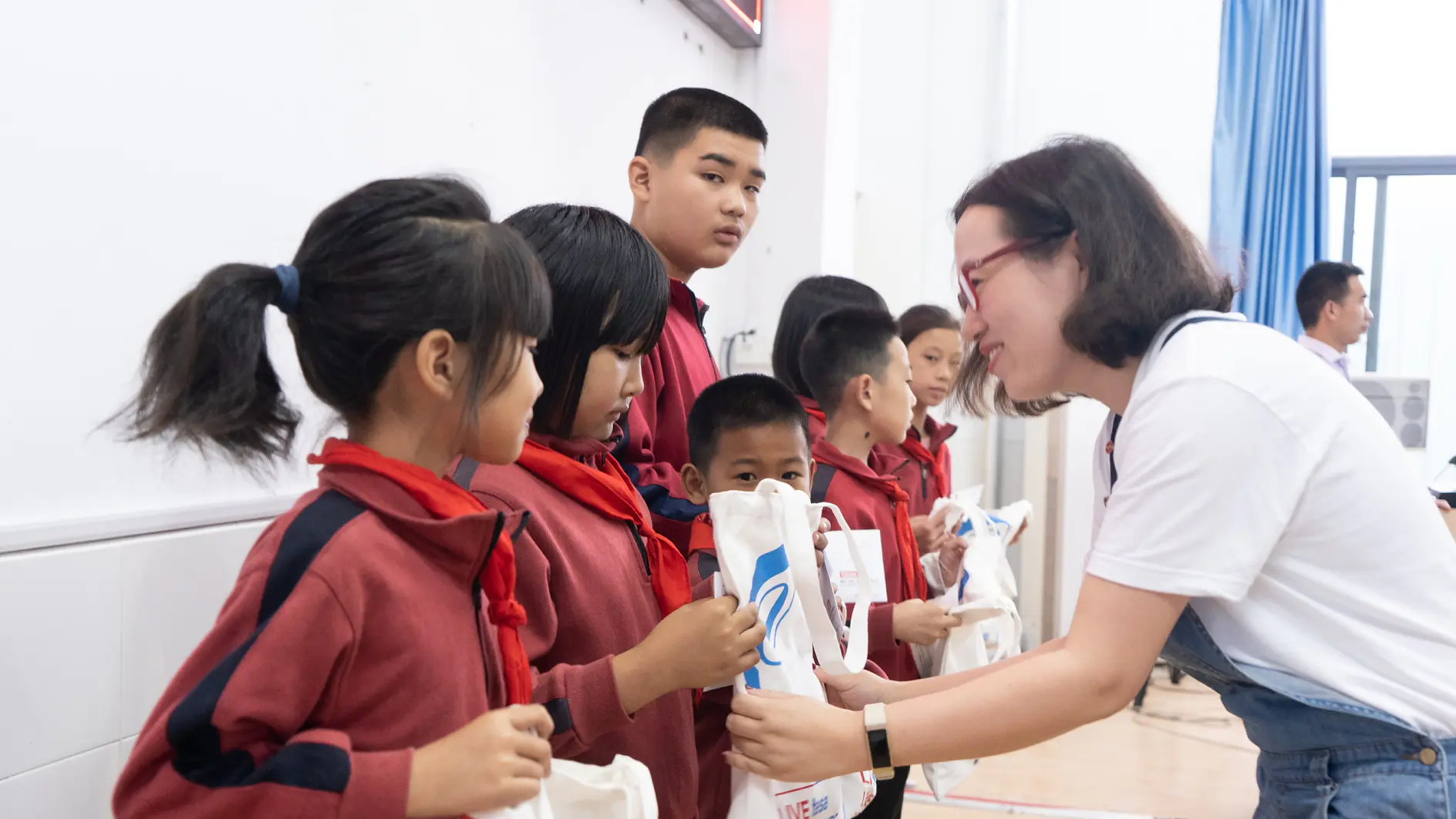 Une femme distribue des sacs-cadeaux à un groupe d’enfants dans une salle de classe. Les enfants sont alignés, portant des uniformes rouges et noirs avec des foulards rouges. La femme utilise du ruban tesa pour sceller l’emballage de chaque sac-cadeau. Elle porte des lunettes et est habillée de façon décontractée.