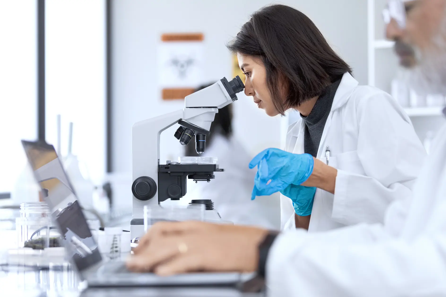 Une femme en blouse de laboratoire utilise un microscope dans un environnement de laboratoire. Elle porte des gants bleus et est très concentrée. Au premier plan, un homme en blouse de laboratoire travaille sur un ordinateur portable. Du matériel scientifique, y compris du ruban tesa, est visible autour d'eux.