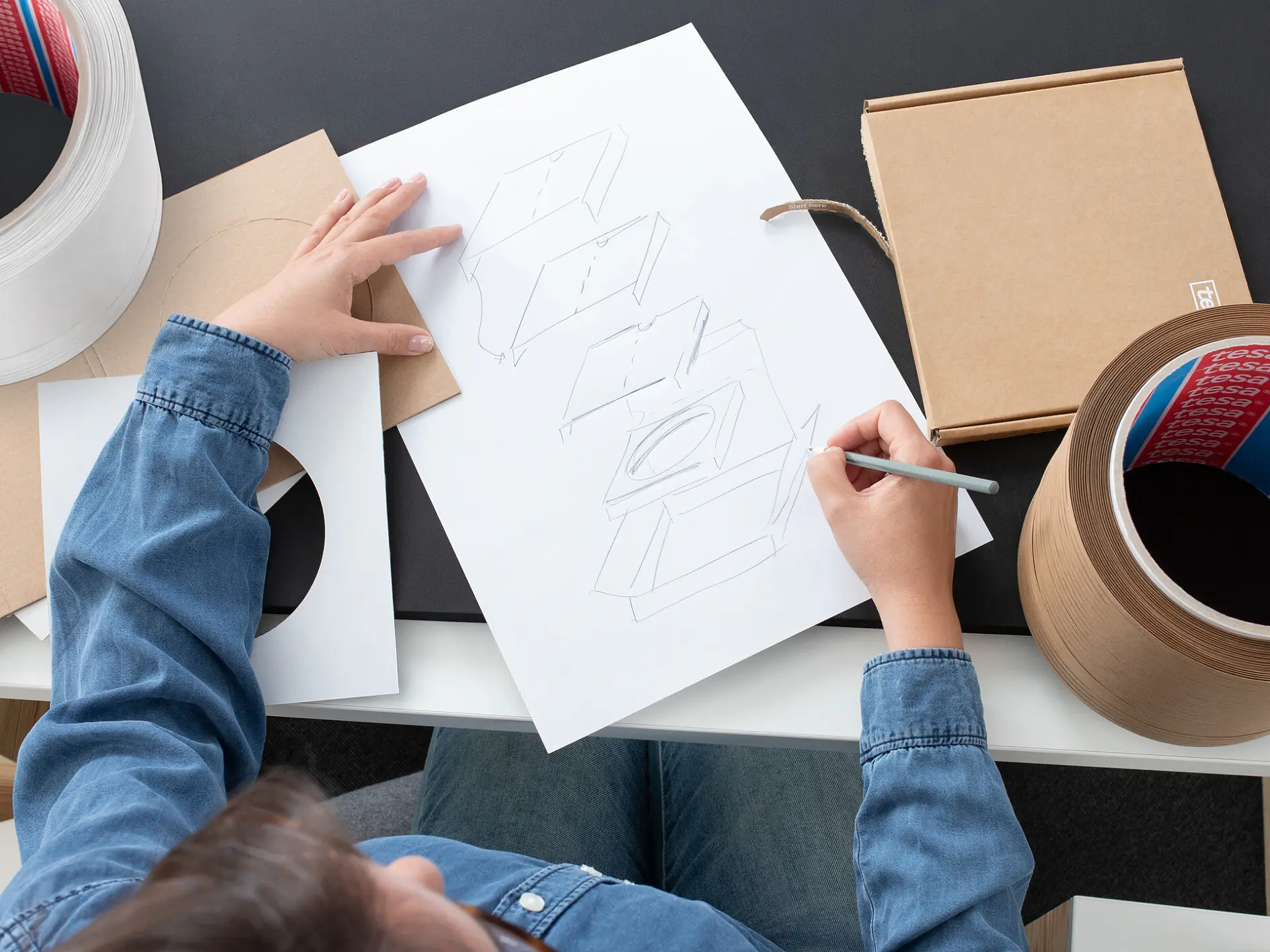 personne esquissant des designs d'emballages de produits sur du papier à un bureau, avec des boîtes en carton et des matériaux d'emballage à proximité