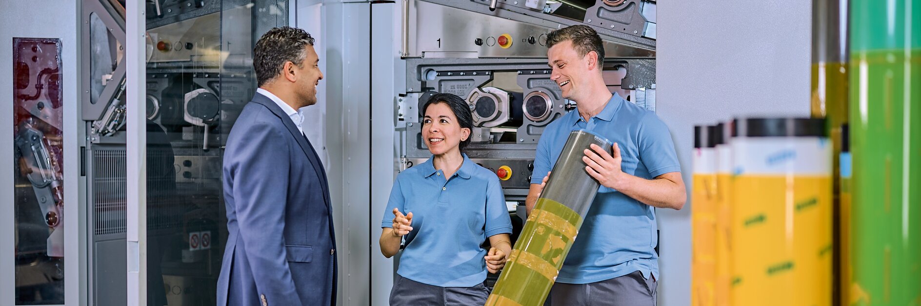 two men and one woman in a factory setting inspecting large rolls of tesa adhesive tape near industrial machinery