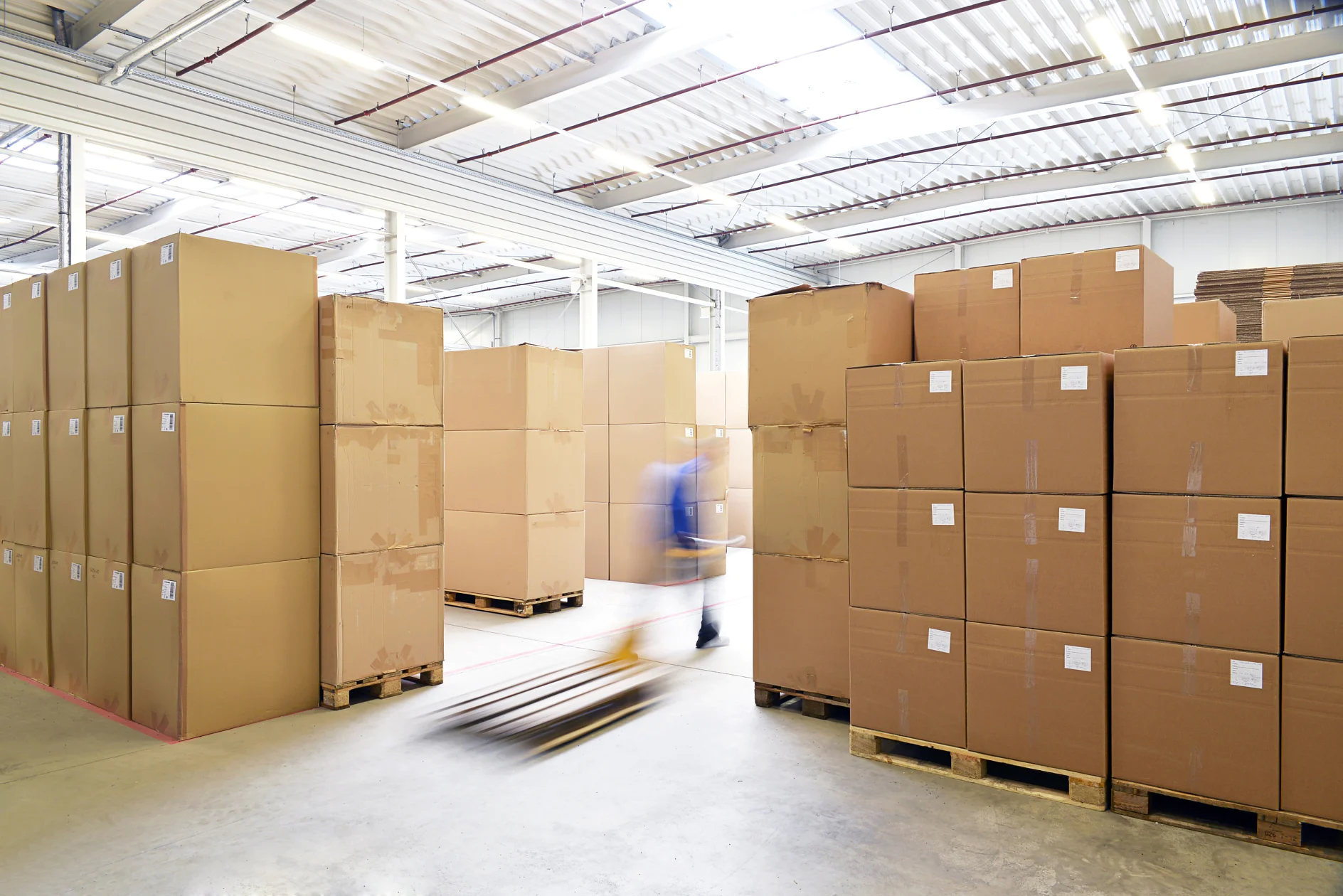 A large warehouse with stacks of cardboard boxes on wooden pallets. A person using a hand pallet truck is moving between the rows. The space is well-lit with natural light coming through the ceiling. The floor is concrete, and tesa tape is used throughout for securing packages and ensuring everything stays in place during transport. (This text has been generated by AI)