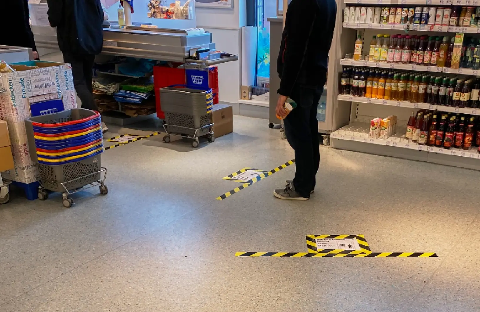 A supermarket scene shows social distancing markers on the floor with yellow and black tesa tape. Two people stand within the marked areas near shelves stocked with beverages and a table of colorful baskets. Shopping carts are nearby. (This text has been generated by AI)