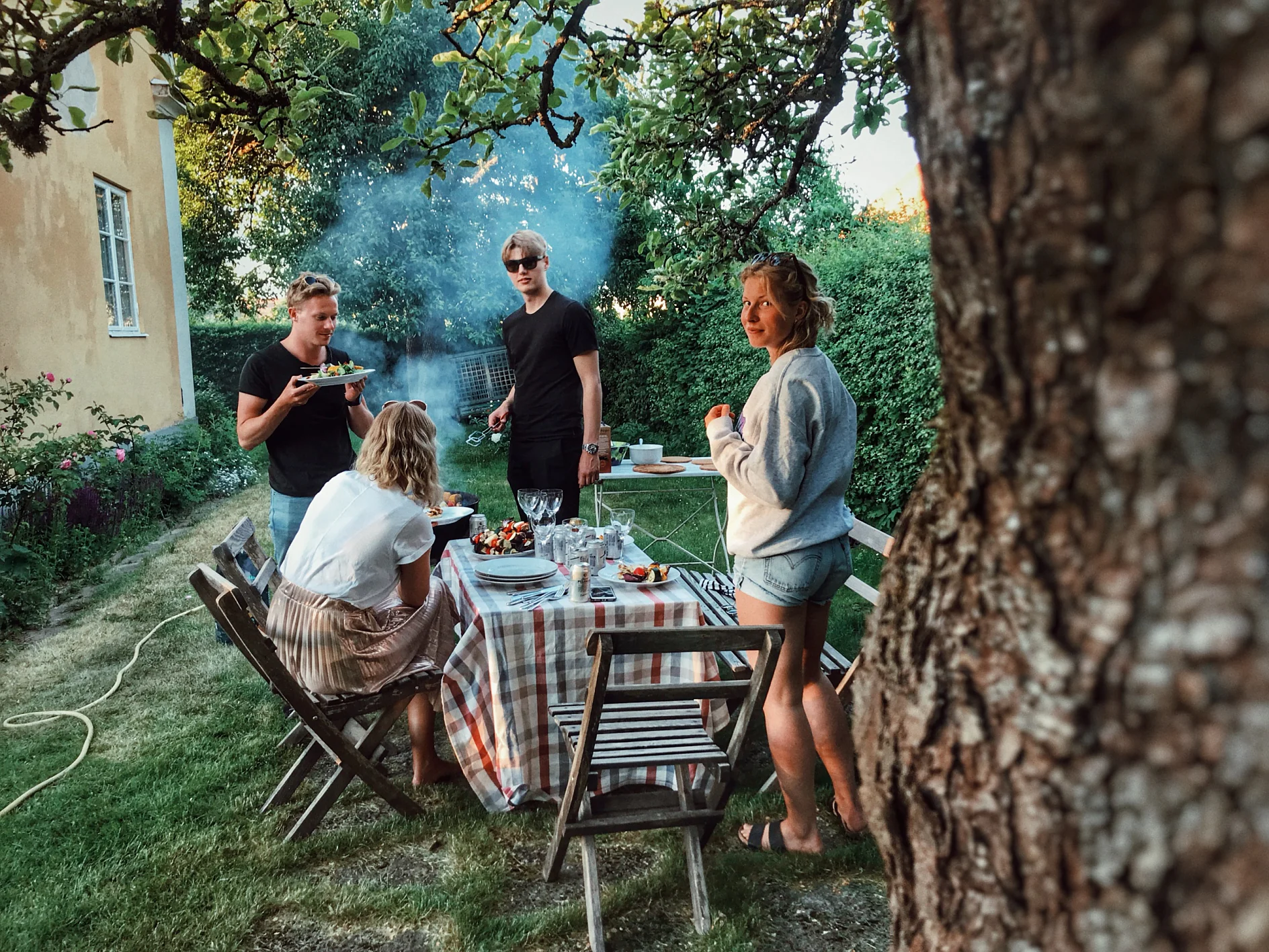 Négy ember gyűlik egy asztal körül egy szabadtéri étkezéshez a kertben. Egy ember tányérból eszik, míg mások állnak és beszélgetnek. Az előtérben egy fa, a háttérben egy grillrácsból száll fel a füst. (This text has been generated by AI)