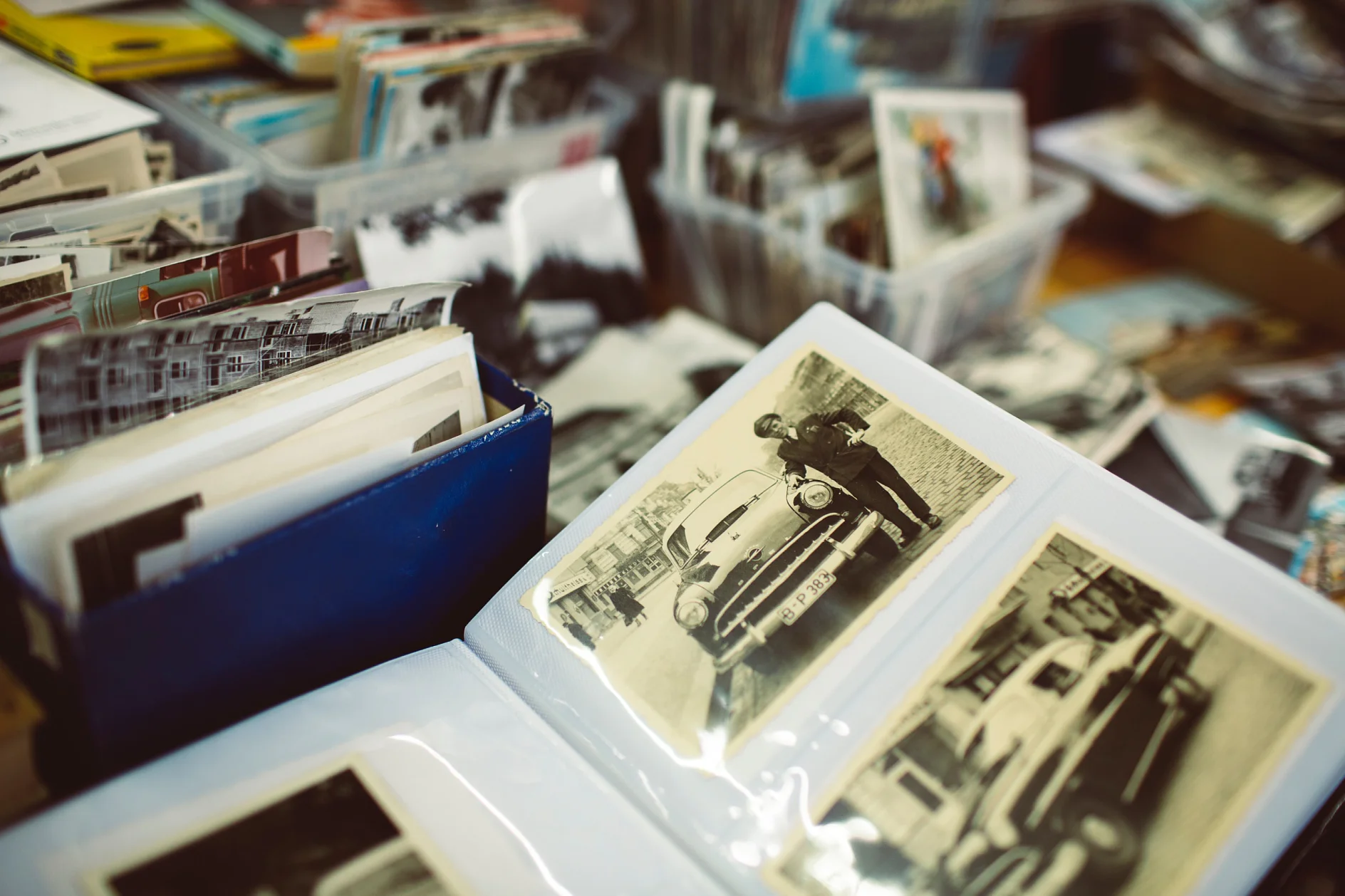 Vintage fényképek gyűjteménye terül el az asztalon. Több fekete-fehér fotó albumba van rendezve, míg másokat műanyag tartályokban tárolnak. Az album egyik fotóján egy személy látható egy klasszikus autó mellett, tesa ragasztószalaggal biztonságosan tartva. (This text has been generated by AI)