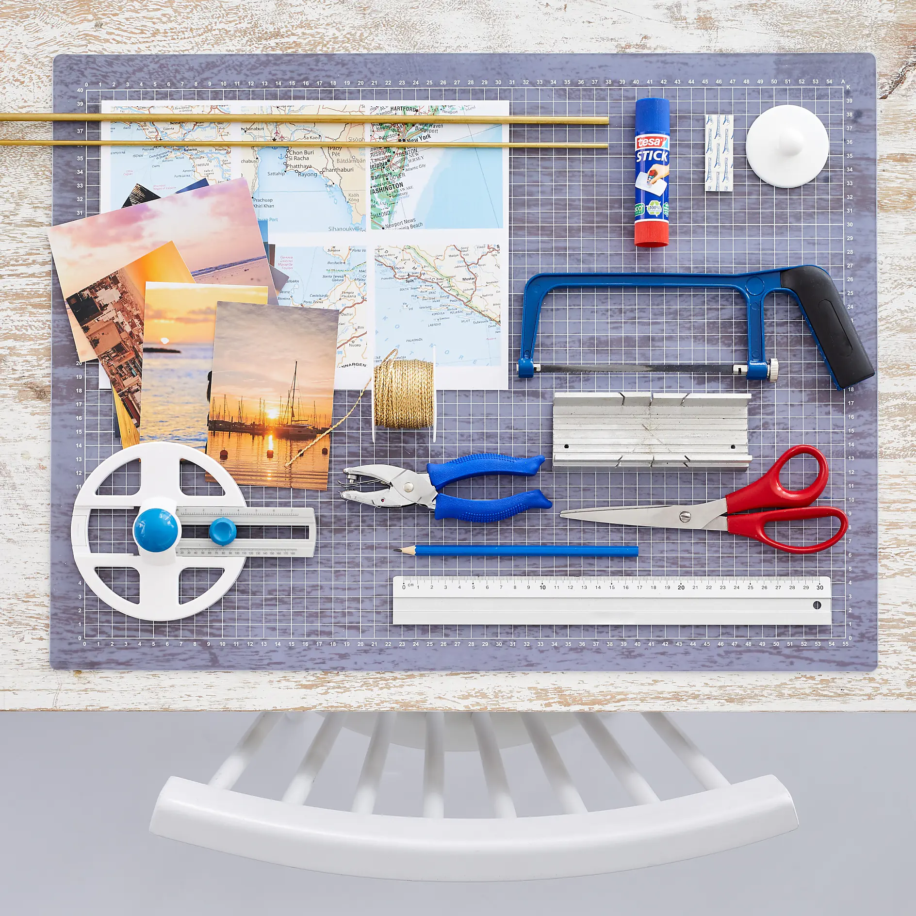 A crafting workspace with maps, sunset photos, a small saw, blue pliers, red scissors, a glue stick, tesa tape, string, a ruler, a circular cutting tool, and a small cutting board on a grid-patterned mat. A white chair is partially visible at the bottom. (This text has been generated by AI)