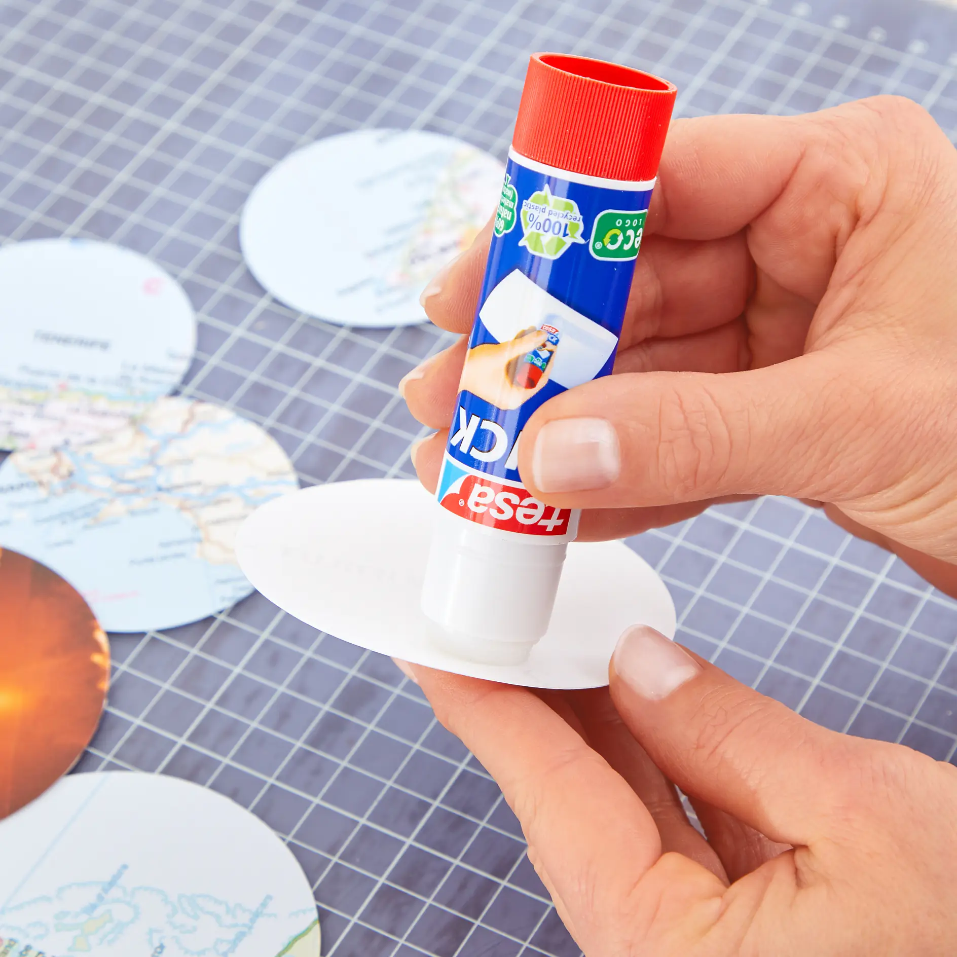 A person applies tesa tape onto a circular piece of paper. Several paper circles with map patterns are on a grid-patterned surface in the background. The tesa tape dispenser has a red cap and a blue label. (This text has been generated by AI)