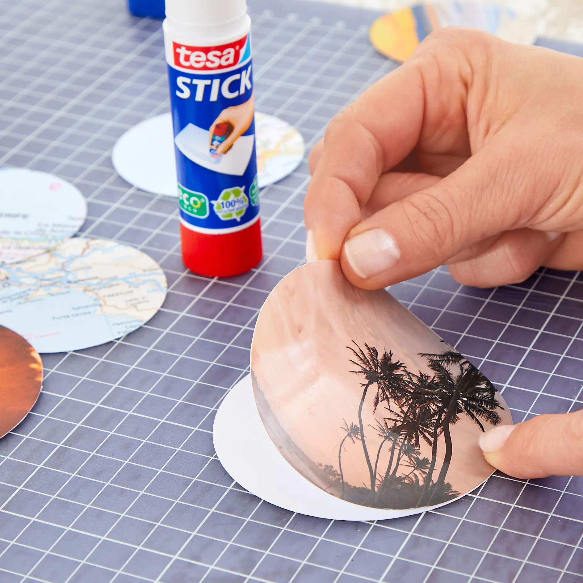 Hand holding a cut-out of a round image with palm trees and a sunset, placing it on white paper. A tesa tape roll is on the grid-patterned surface, along with other circular cut-outs showing maps and images. (This text has been generated by AI)