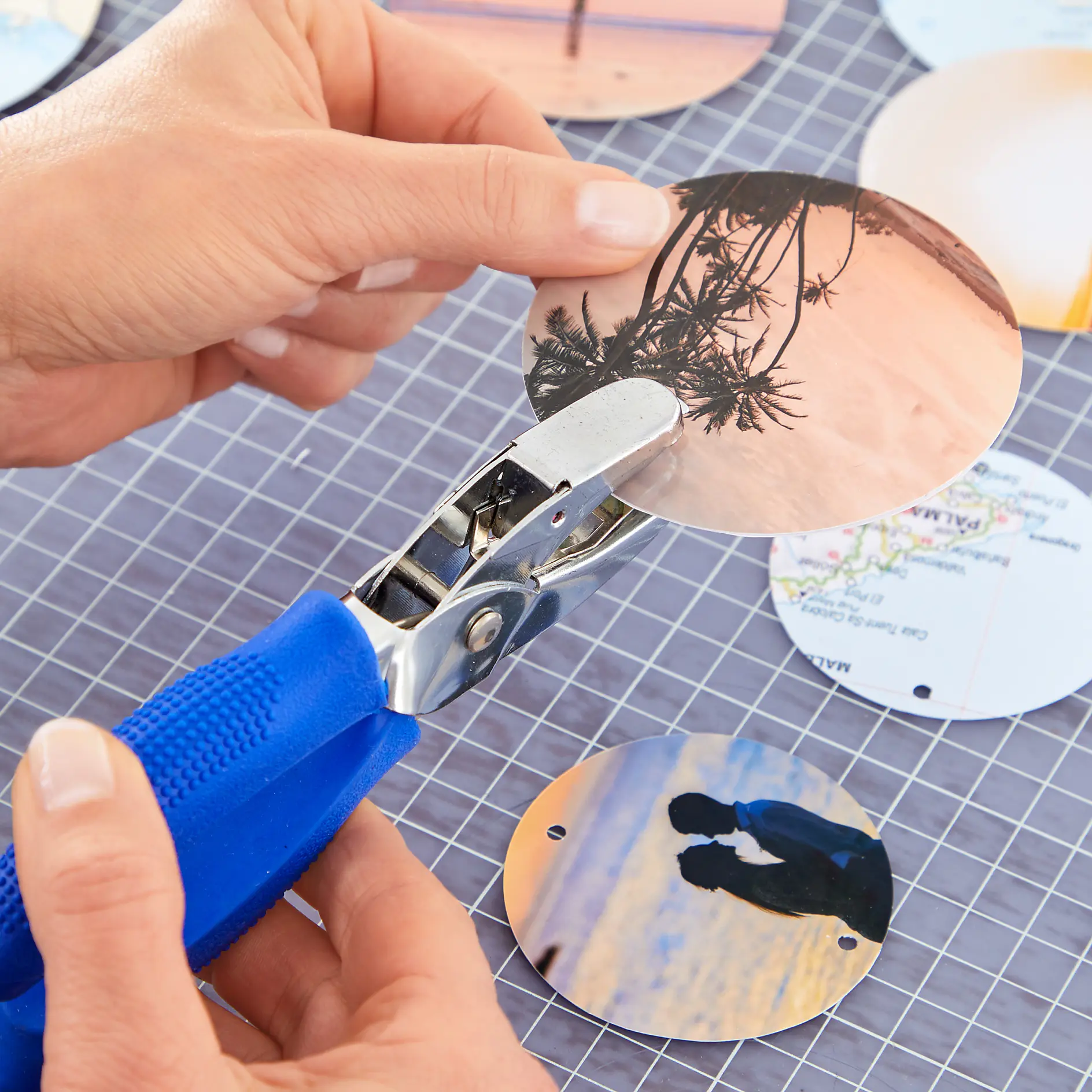 A person uses a blue-handled hole punch to make a hole in a circular photo of a beach scene. Other circular photos with holes are on a grid-patterned surface held in place with tesa tape. (This text has been generated by AI)