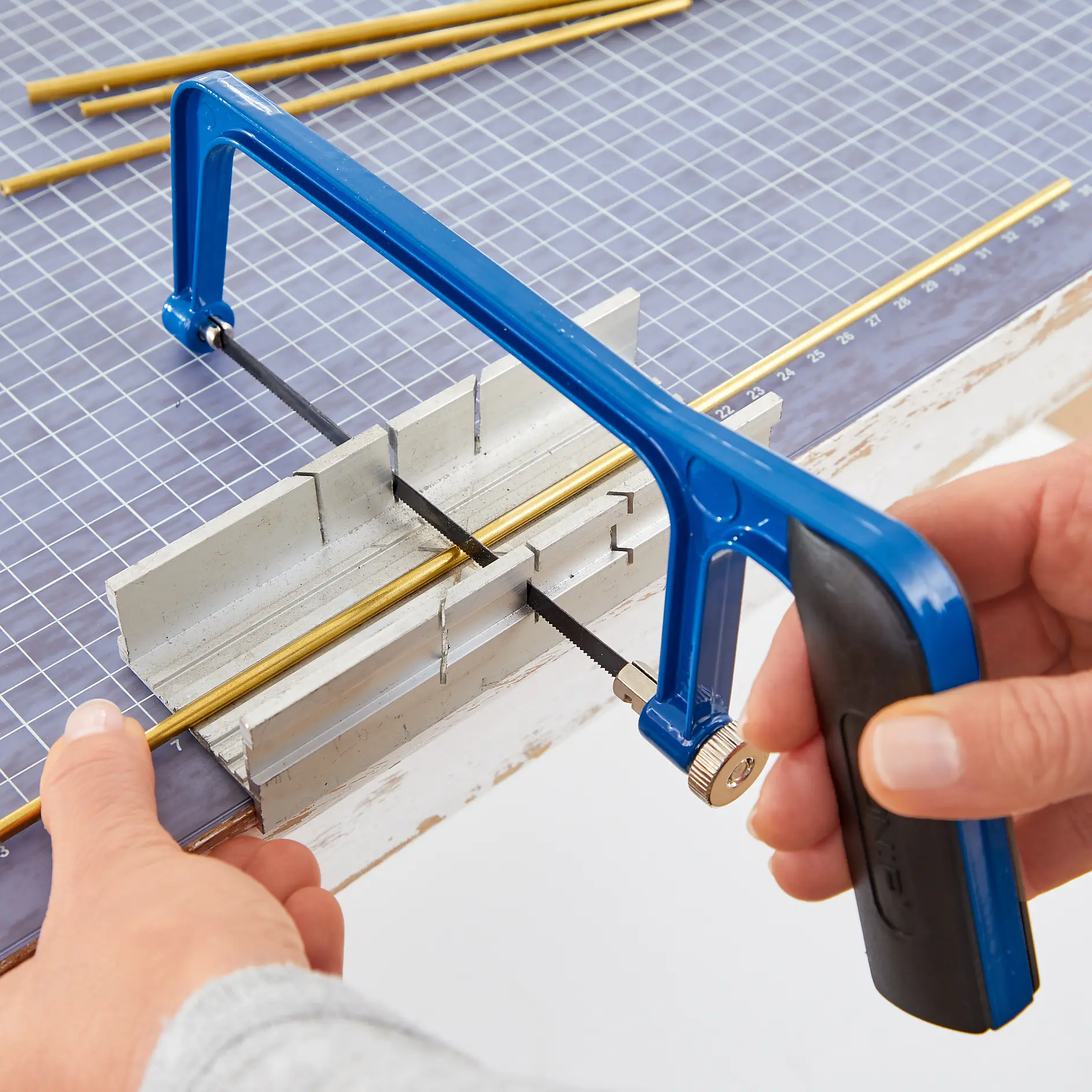 A person uses a blue hacksaw to cut a thin brass rod on a miter box. The background shows a workspace with a grid-patterned surface, additional rods, and tesa tape for measuring. (This text has been generated by AI)