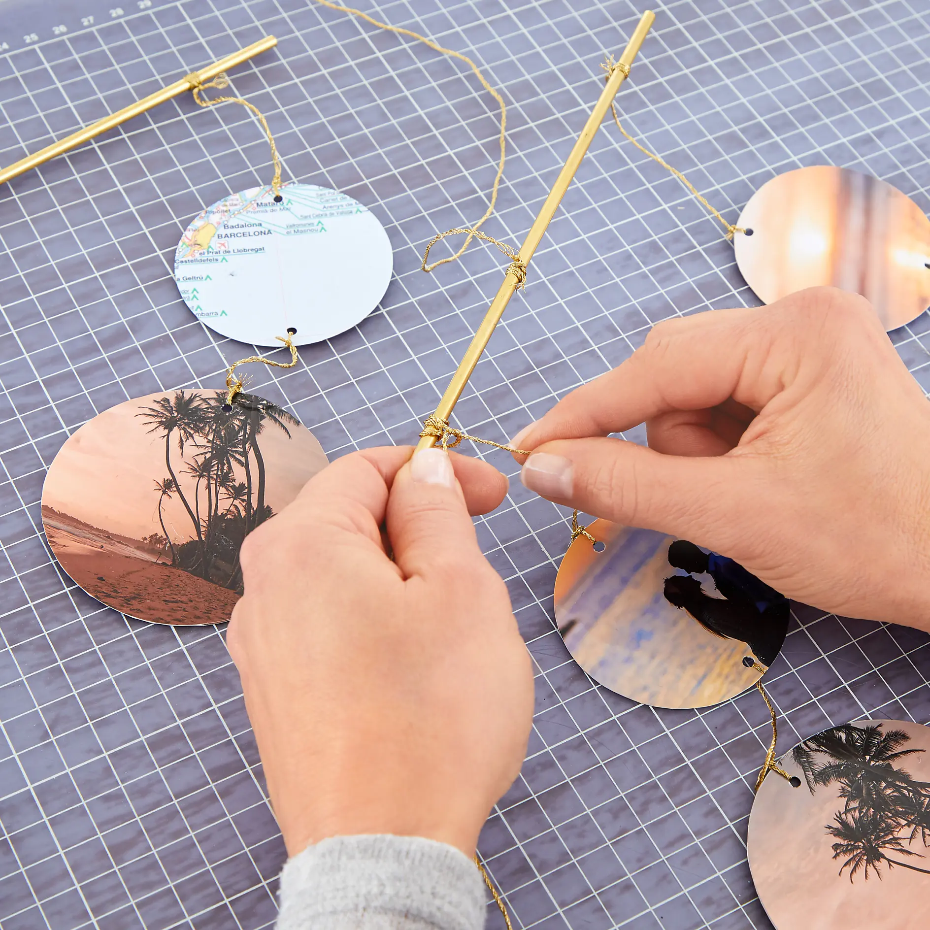 Hands are seen tying string to a circular photo print, assembling a hanging decor piece. Several other circular prints are already attached using tesa tape. A grid-lined mat is underneath, and a thin, gold-colored rod is used as the frame. (This text has been generated by AI)