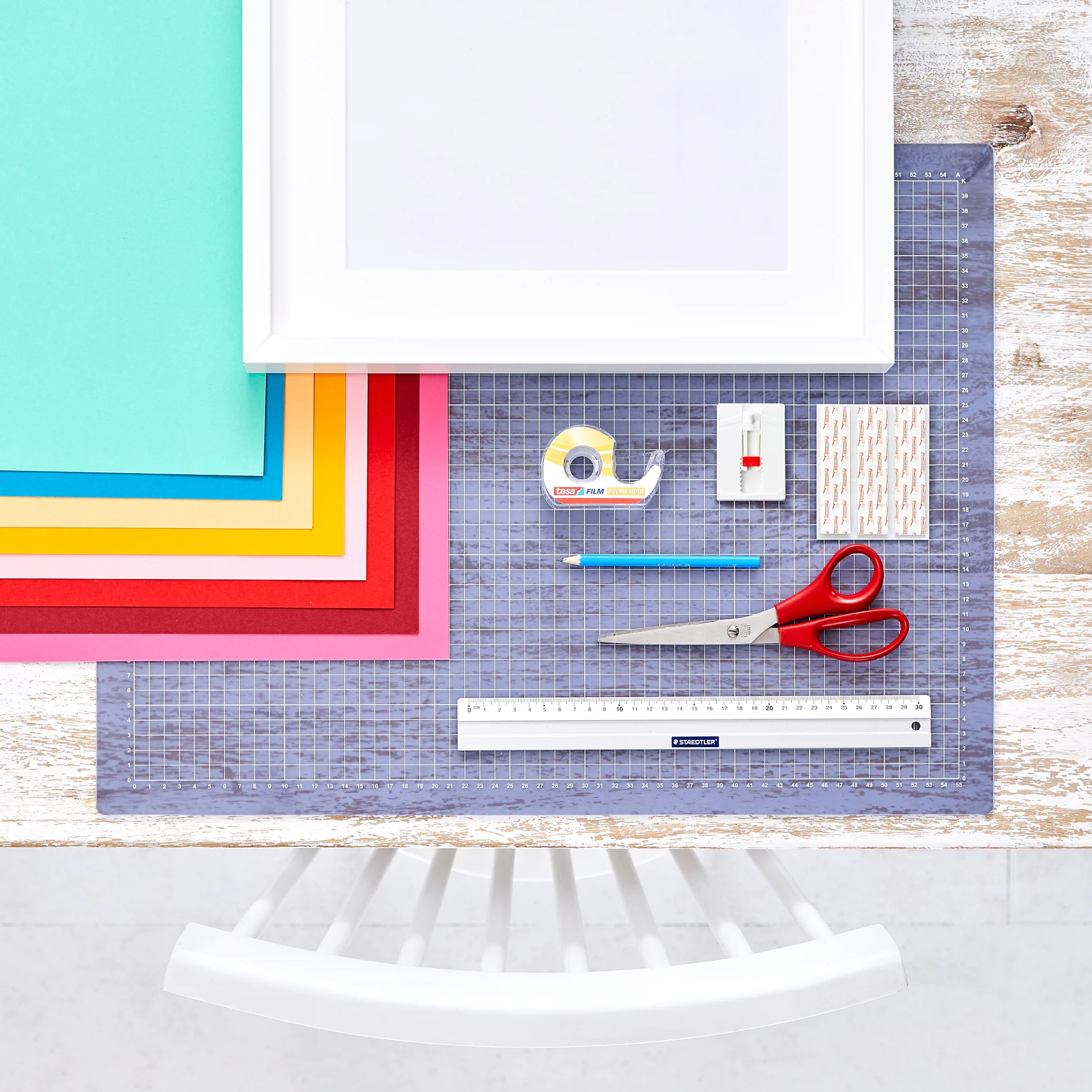Rendben elrendezett lapos kompozíció egy világos faasztalon: egymásra rakott színes papírok, vonalzó, olló, ceruza, tesa ragasztószalag, hegyező és ragasztócsíkok. Alul részben látszik egy fehér szék. (This text has been generated by AI)