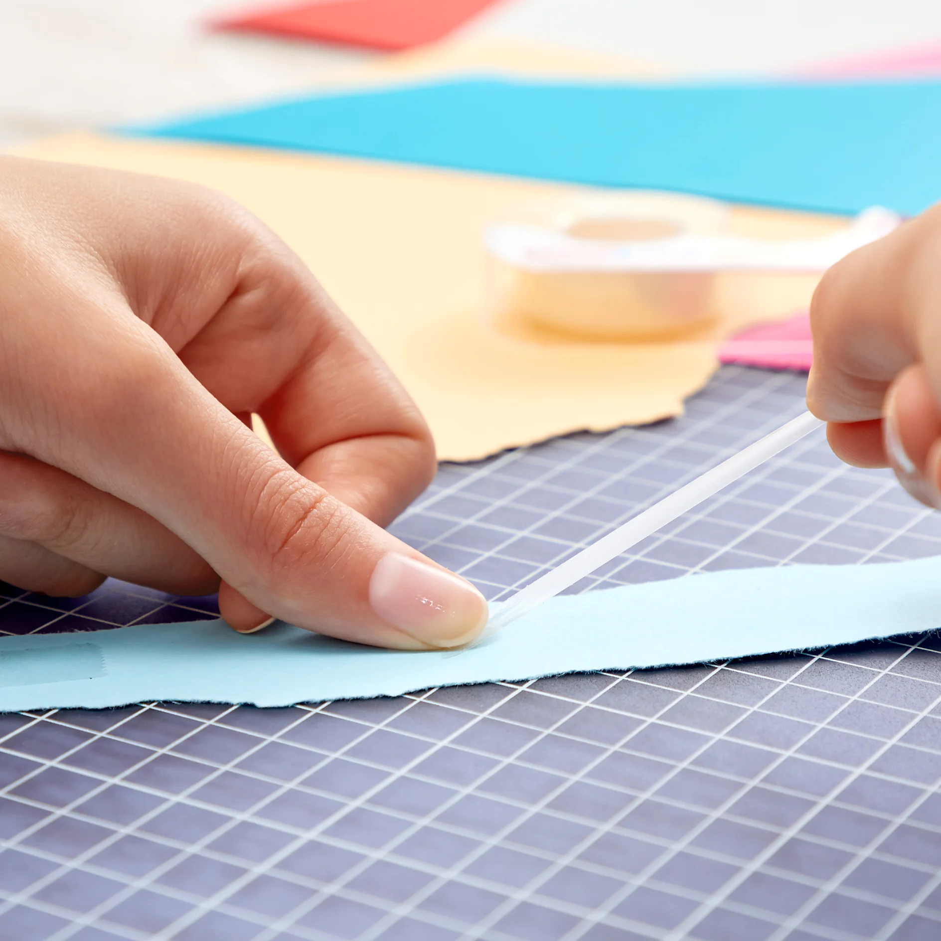 Kézek egy csík tesa ragasztószalagot helyeznek egy kék papírra, rácsmintás vágólapon. A háttérben színes papírok és tesa szalag látható. (This text has been generated by AI)