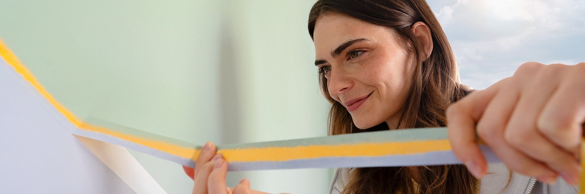 A woman is applying tesa tape to a wall with a light green color. She appears focused on ensuring the tesa tape is straight. She has long brown hair and is wearing a light-colored hoodie. There is natural light coming through a window. (This text has been generated by AI)