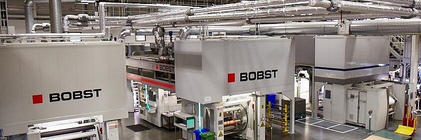 A large industrial facility with multiple Bobst machines for printing or packaging. The machines are white with red and black logos. The room has a high ceiling with exposed ducts and overhead lighting. Various materials, including tesa tape, and bins are visible. (This text has been generated by AI)