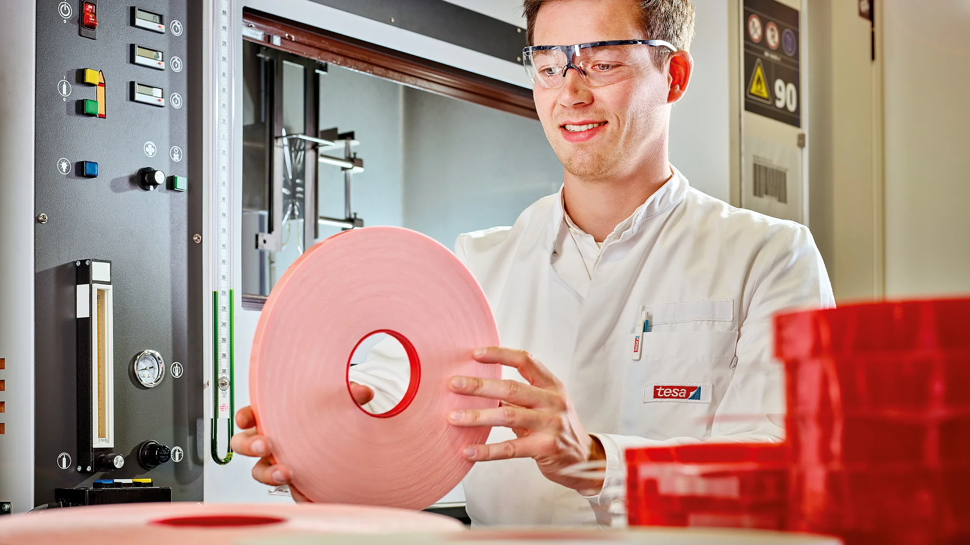 A person wearing safety glasses and a white lab coat holds a large roll of pink tesa tape. Various control panels and rolls of red tesa tape are visible in the background. (This text has been generated by AI)
