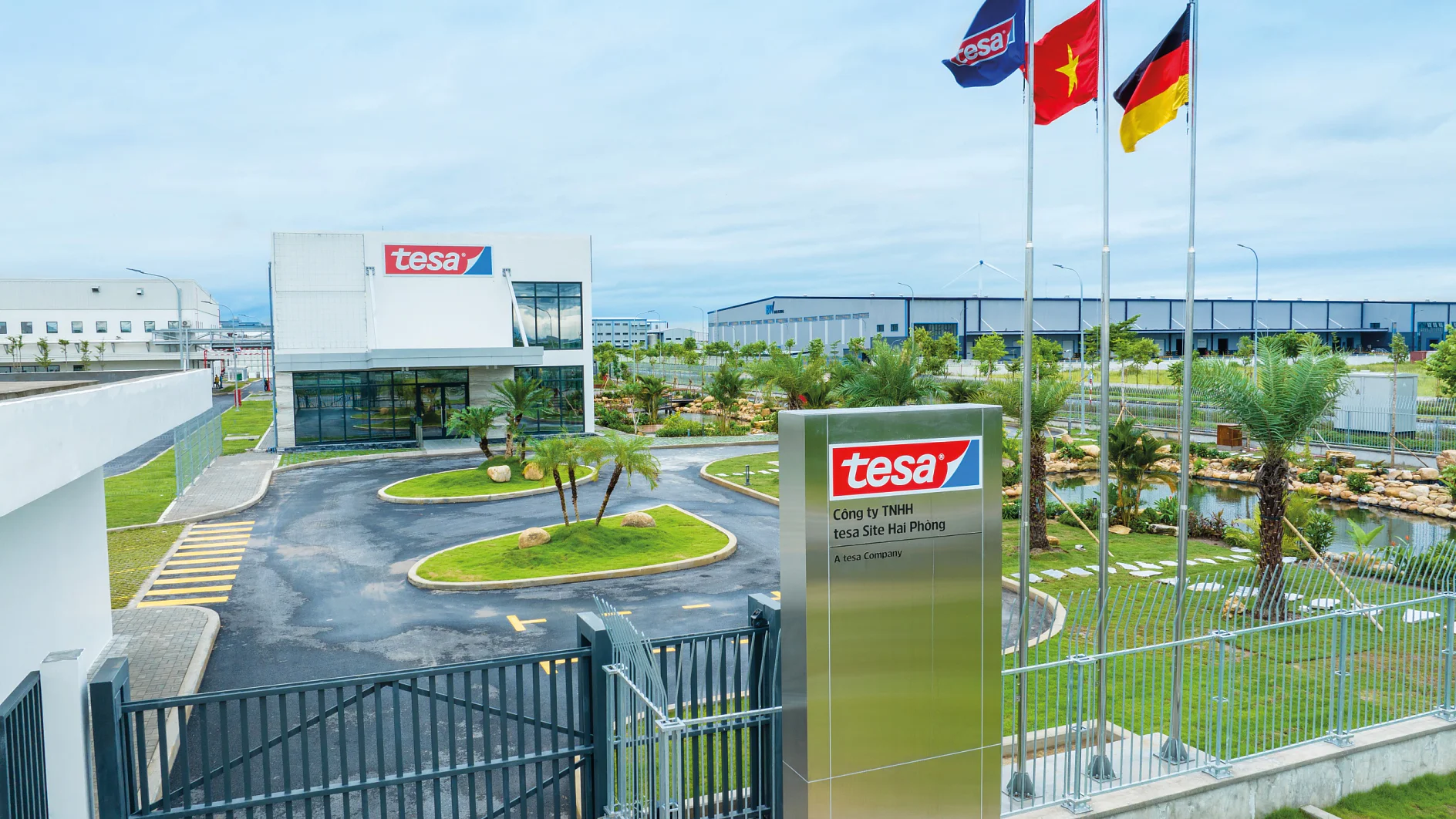 Entrance of a modern industrial facility with a sign reading tesa. Two buildings feature the tesa logo. Three flags fly near the entrance. The landscaped area includes trees and shrubs, and a roadway leads to the main entrance, prominently showcasing tesa tape throughout. (This text has been generated by AI)