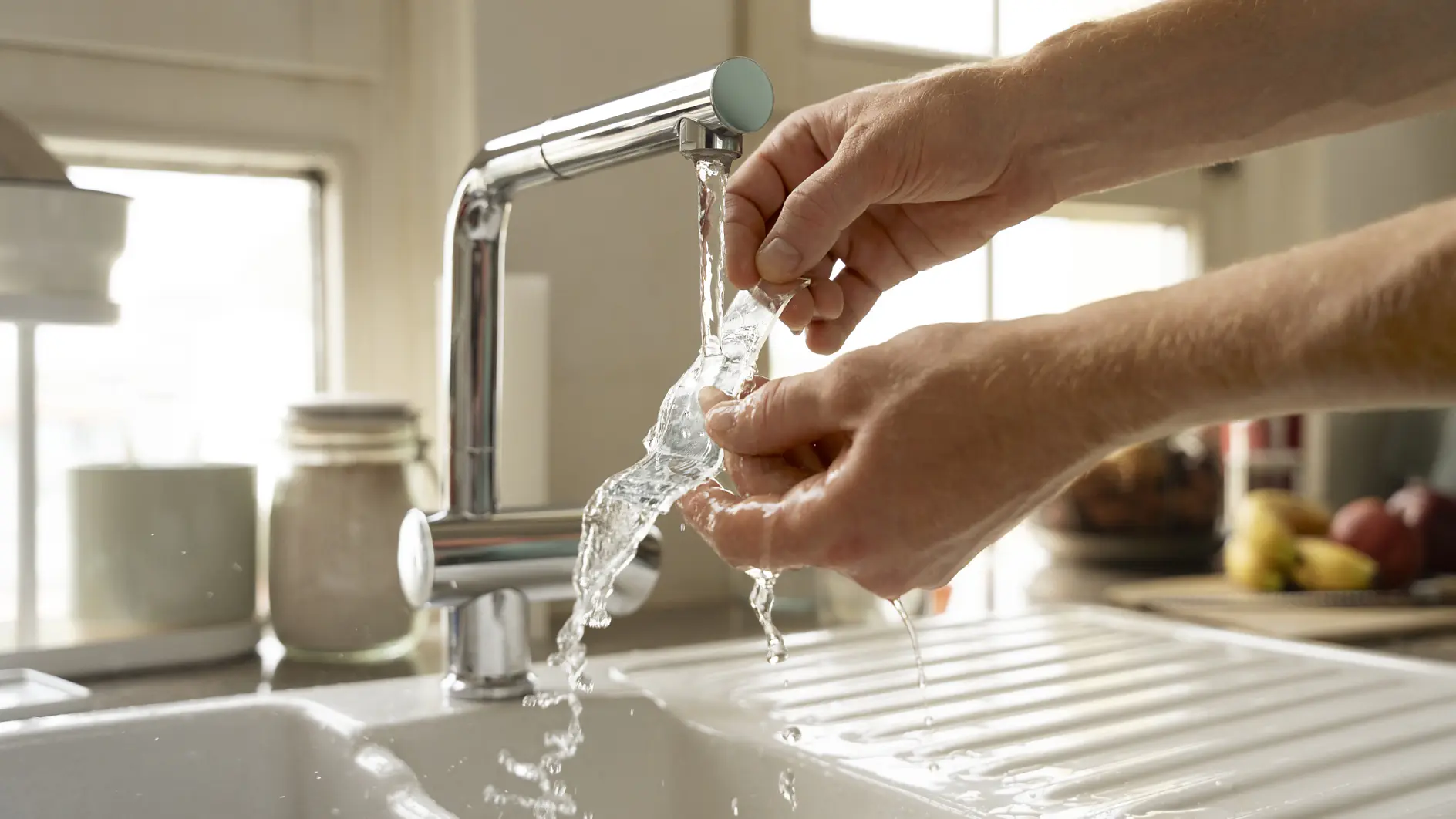 Hands are rinsing a transparent plastic bag under a running kitchen faucet. Water splashes into the white sink below, with glass jars and fruit visible in the background. (This text has been generated by AI)