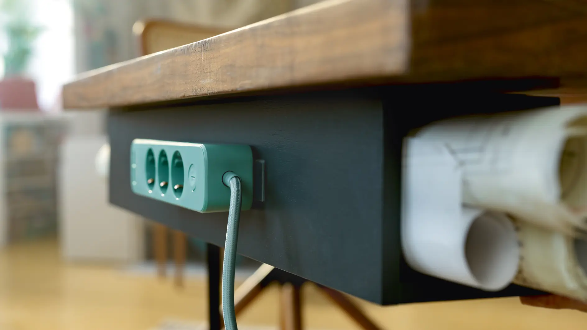 A teal power strip with a braided cable is mounted under a dark wooden desk. Several rolled-up papers are neatly stored on a rack underneath, secured by tesa tape. The background features a chair and a blurred interior setting. (This text has been generated by AI)