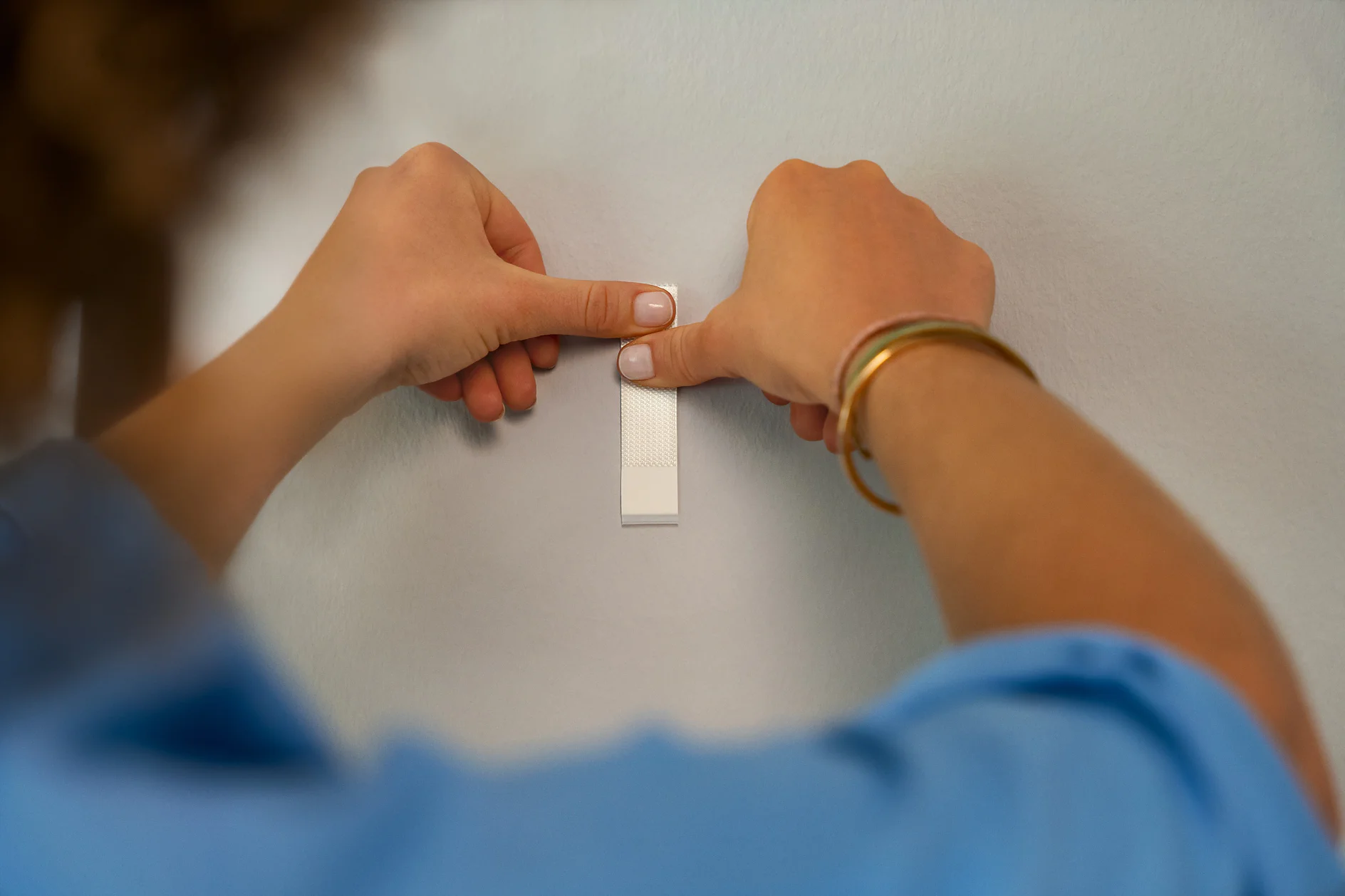 A person in a blue shirt attaches a white tesa tape strip to a white wall using both hands. Their left wrist has a gold bracelet. The image focuses on the hands and the strip. (This text has been generated by AI)