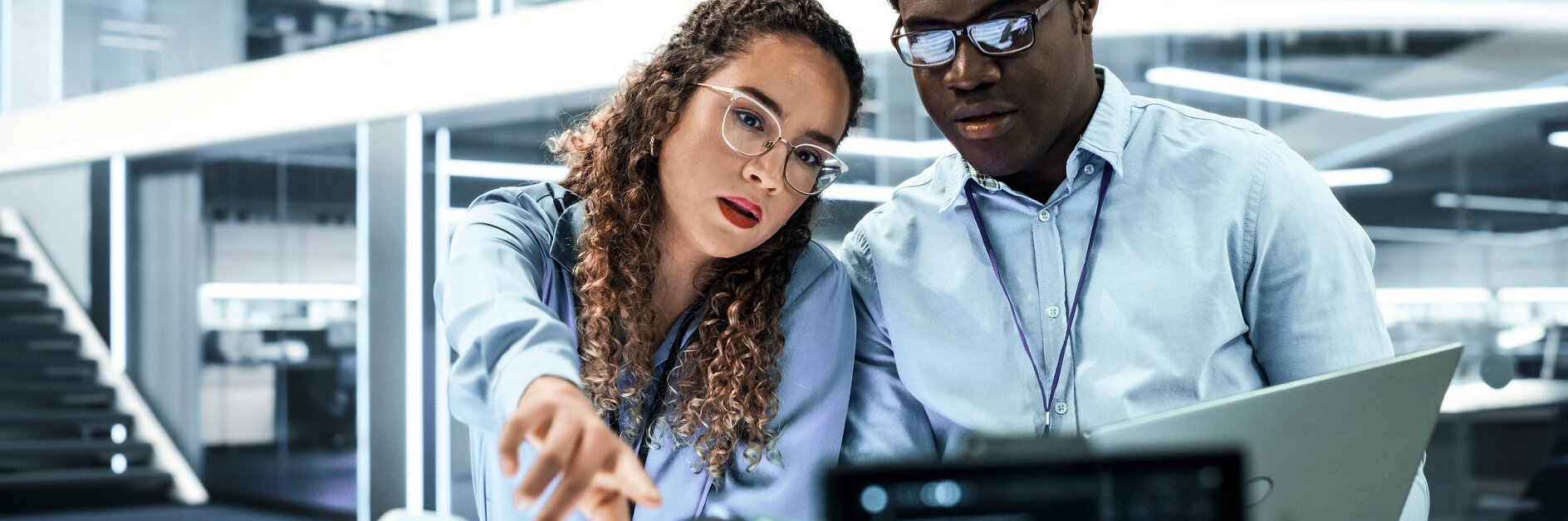 Two professionals in an office discuss data on a laptop and tablet, surrounded by modern glass walls and office equipment.