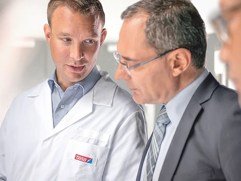 A man in a white lab coat with a tesa tape label talks to another man in a suit and glasses. They appear to be in a discussion while looking at something out of the frame. The background is bright and slightly blurred. (This text has been generated by AI)