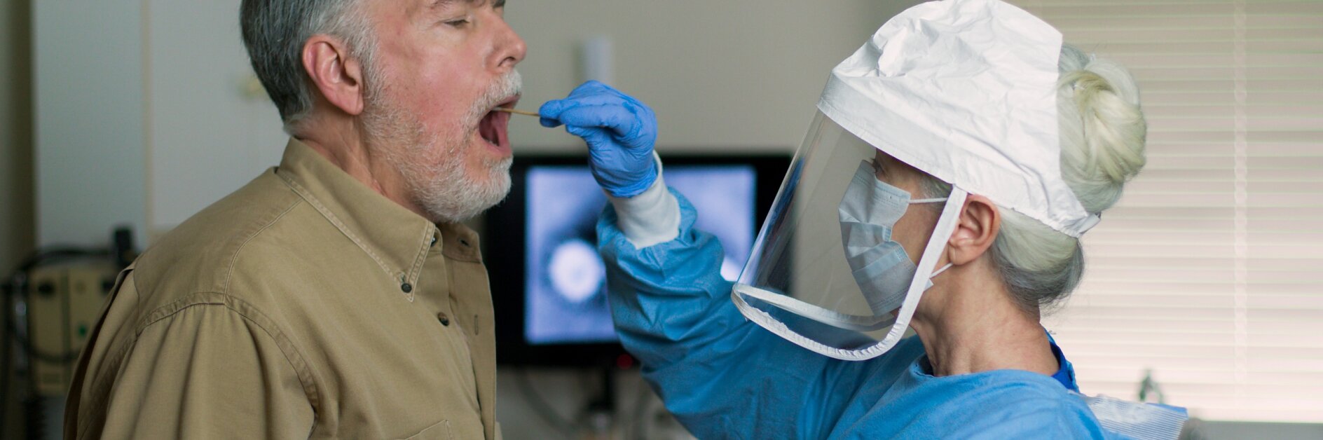 A medical professional in protective gear, including a face shield and mask, collects a nasal swab sample from an older man in a clinical setting. The man is standing with his mouth open, and a computer screen is visible in the background. (This text has been generated by AI)