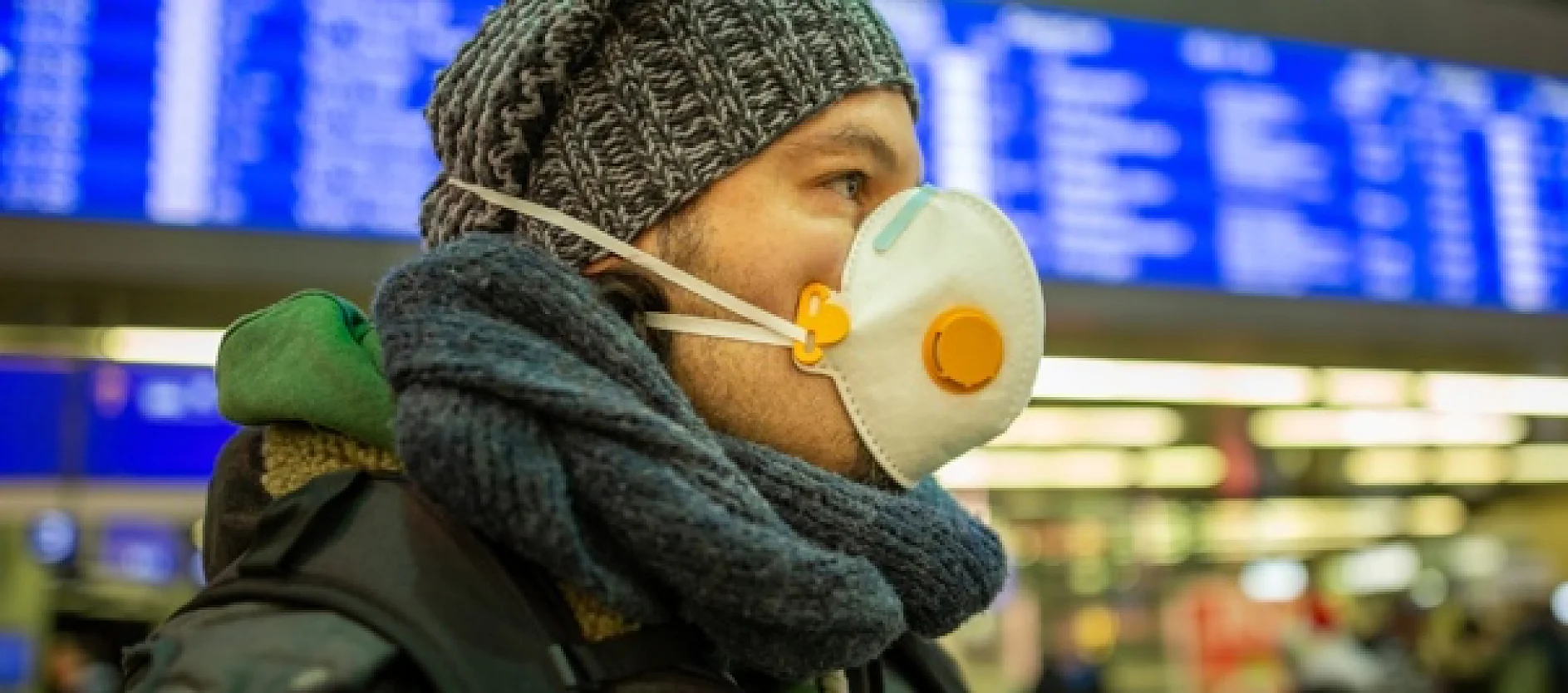 A person wearing a winter hat, scarf, and a protective face mask is in a busy indoor area. Out-of-focus, there are blue information screens displaying departure or arrival details in the background. (This text has been generated by AI)