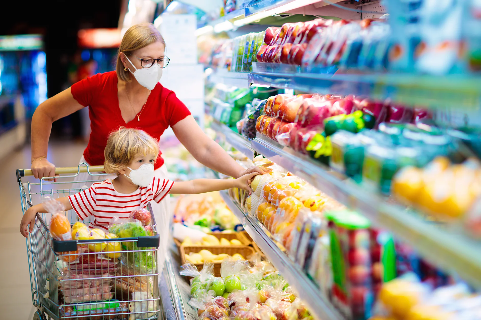 An adult and a child wearing masks are shopping in a grocery store. The child, sitting in a cart filled with fruits, reaches for produce on a shelf. The adult, in a red shirt, is pushing the cart and selecting items. (This text has been generated by AI)