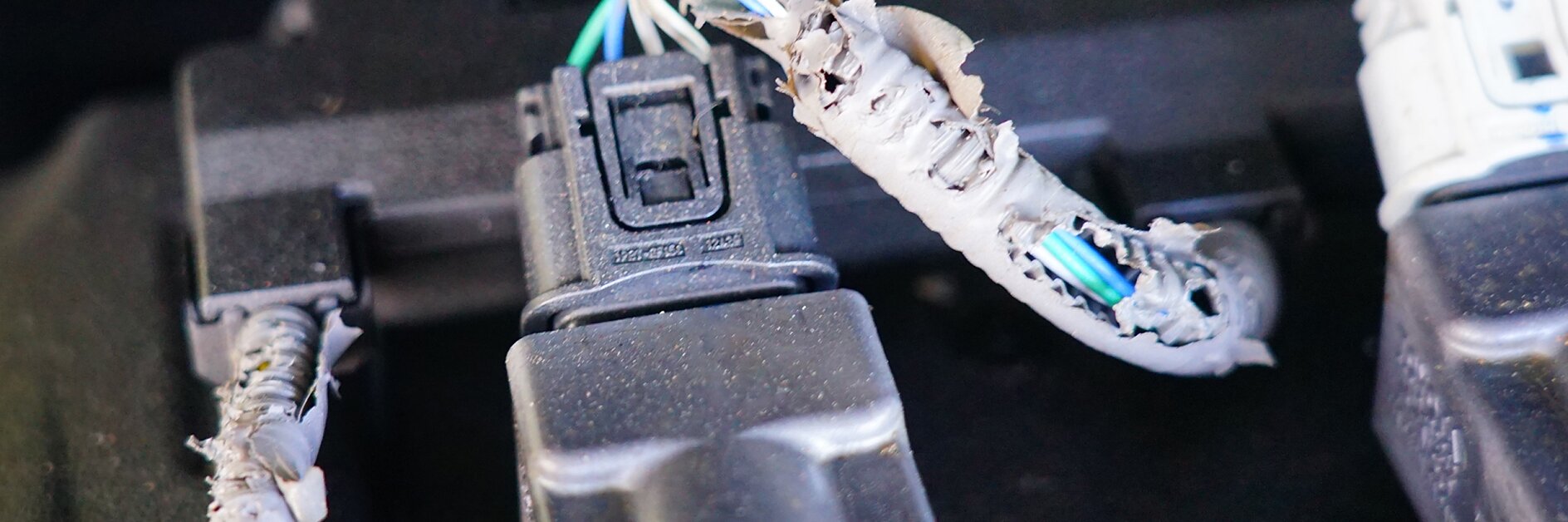 Three electrical connectors with visible damage to the insulation, exposing wires. The insulation appears chewed or torn, showing multiple colored wires inside. The connectors are attached to a black base and wrapped with tesa tape for added protection. (This text has been generated by AI)