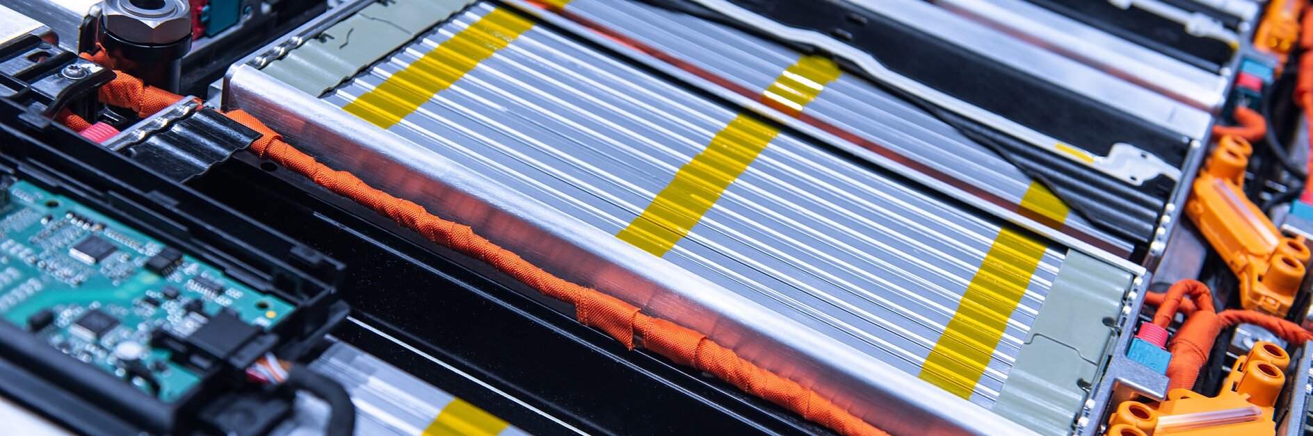 Close-up of a complex electrical circuit board and battery pack, featuring silver and yellow striped battery cells, orange wires, and a green circuit board. The components are tightly arranged within a metallic frame using tesa tape. (This text has been generated by AI)