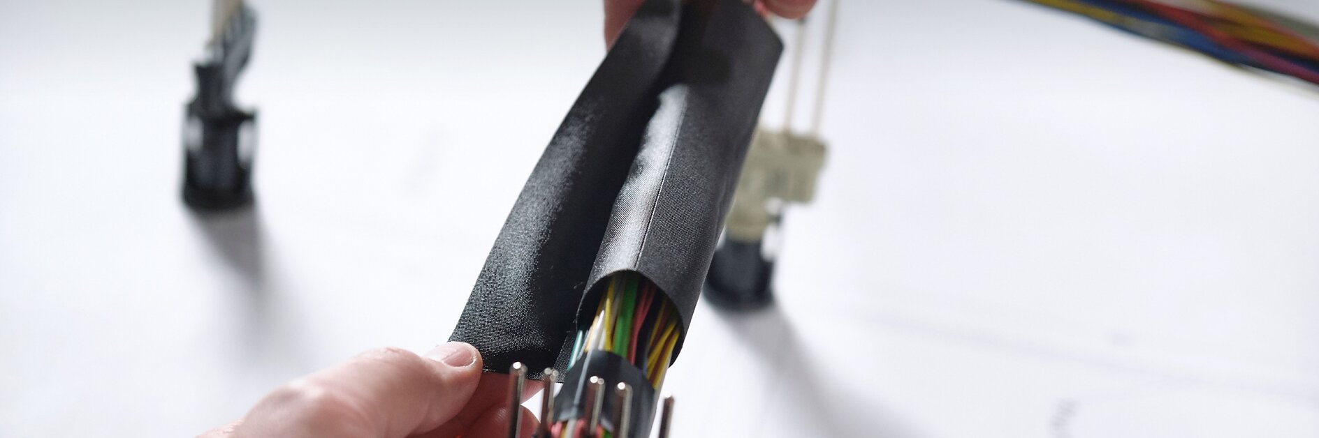 Close-up of hands wrapping a bundle of multicolored electrical wires with black insulating tesa tape. The wires are organized and connected to plastic connectors. A blurred background shows more wires and a surface with printed text. (This text has been generated by AI)