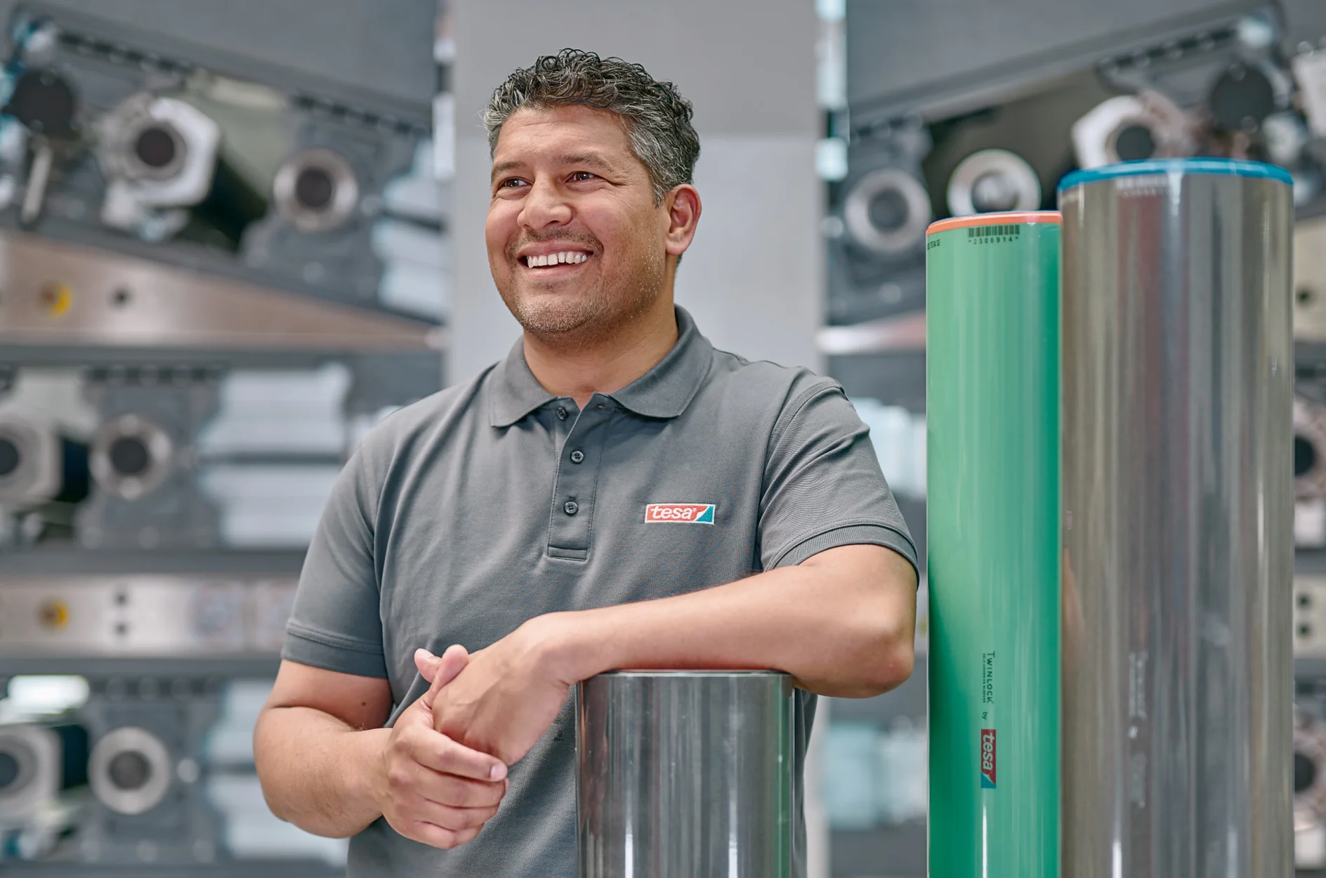 A man in a gray polo shirt stands smiling with his hands resting on a metallic cylinder in a machinery-filled environment. Two large cylindrical rolls, one metallic and one green, stand upright beside him. (This text has been generated by AI)