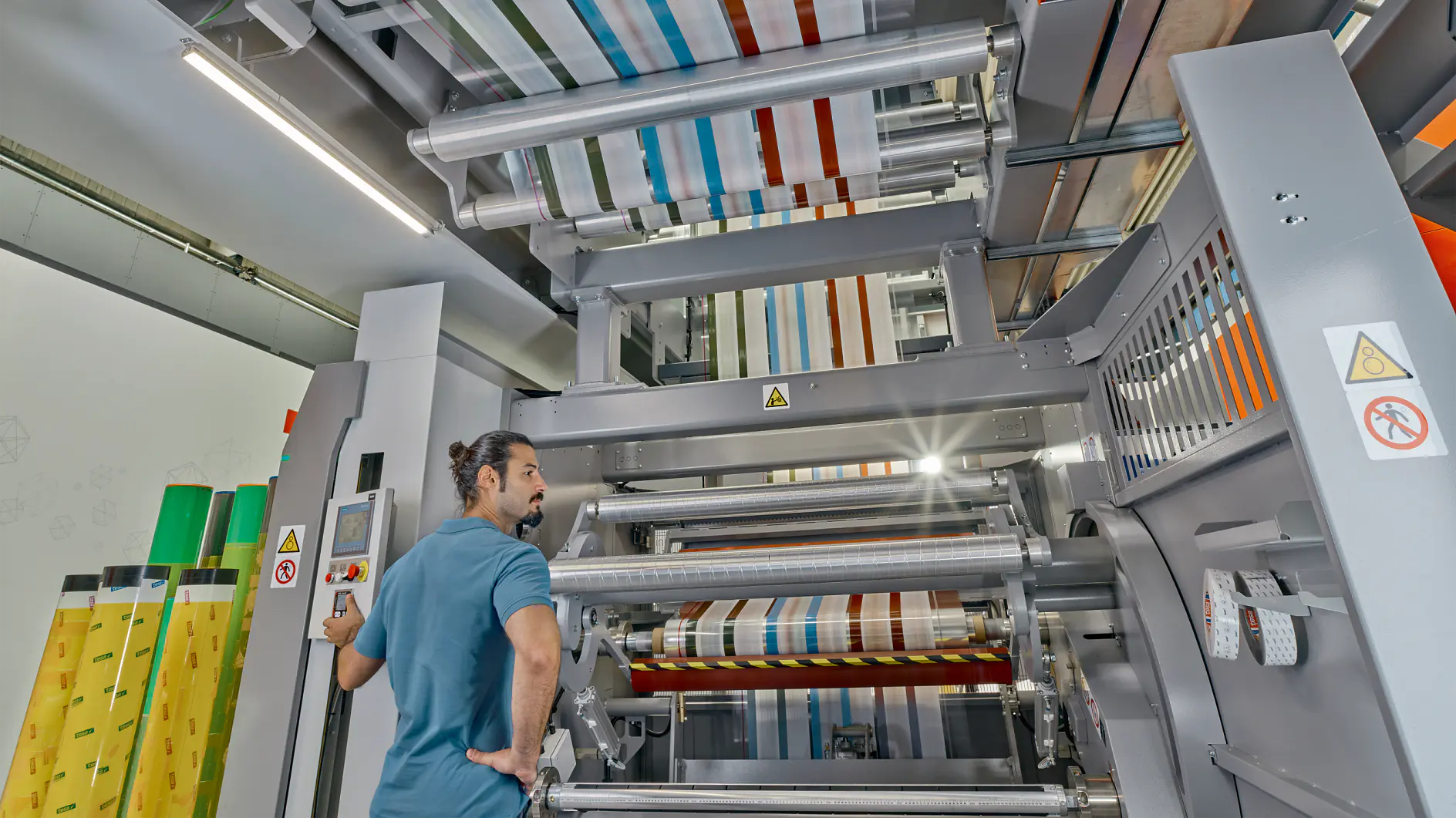 A man with a bun stands near a large industrial machine with multiple rollers, processing colorful striped material wrapped in tesa tape. The machine is metallic and has warning signs. The setting appears to be a factory or manufacturing facility.