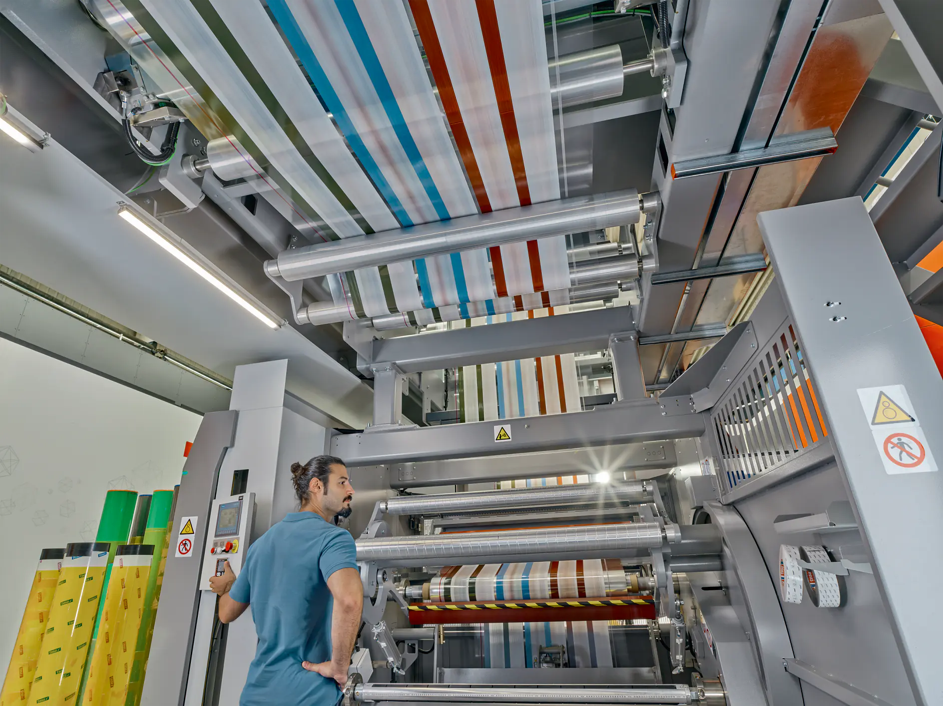 A man with a bun stands near a large industrial machine with multiple rollers, processing colorful striped material wrapped in tesa tape. The machine is metallic and has warning signs. The setting appears to be a factory or manufacturing facility.