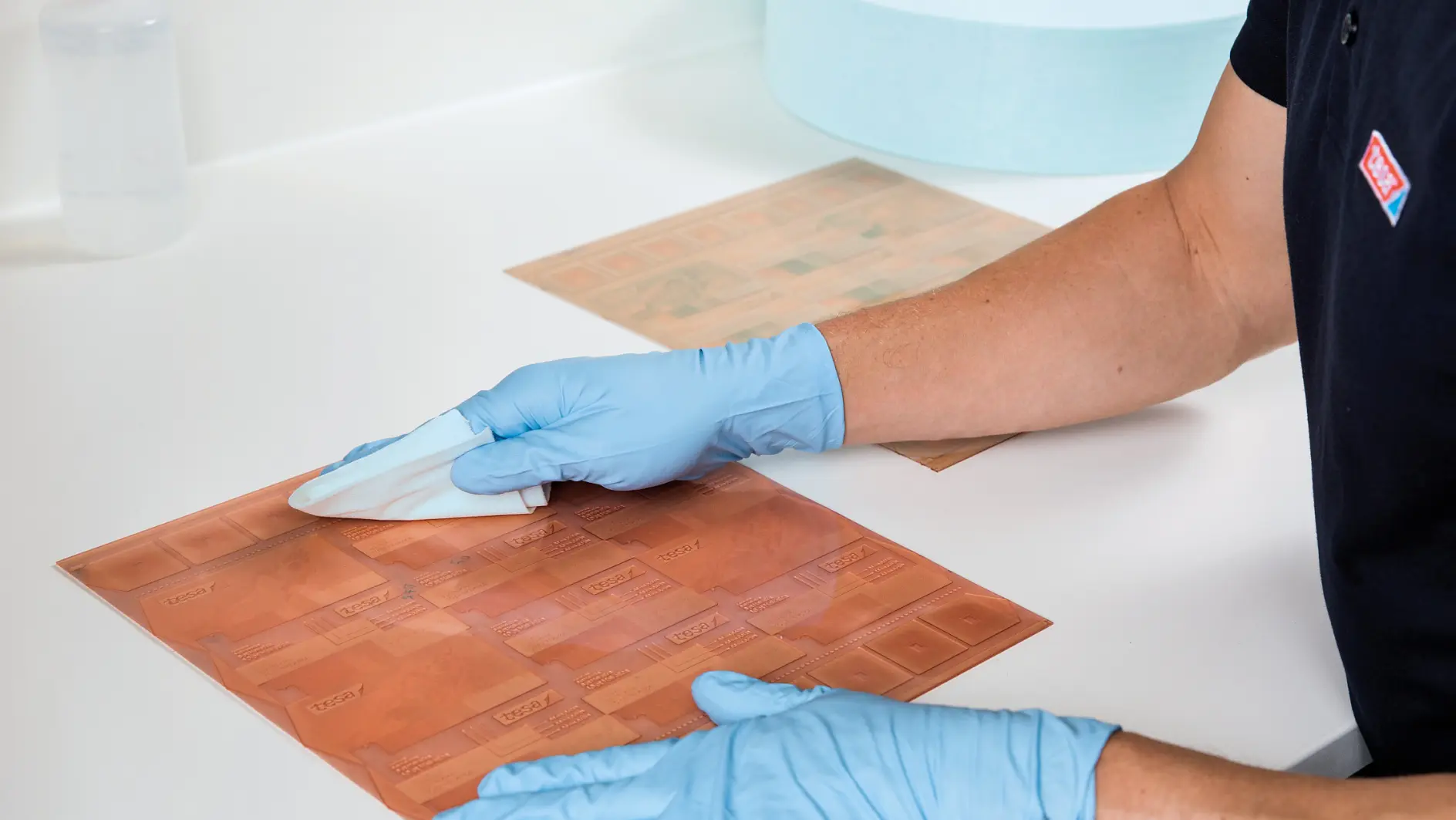 A person wearing blue gloves cleans a rectangular copper plate on a white countertop using a cloth. A bottle of liquid and a large roll of blue tesa tape are visible in the background.