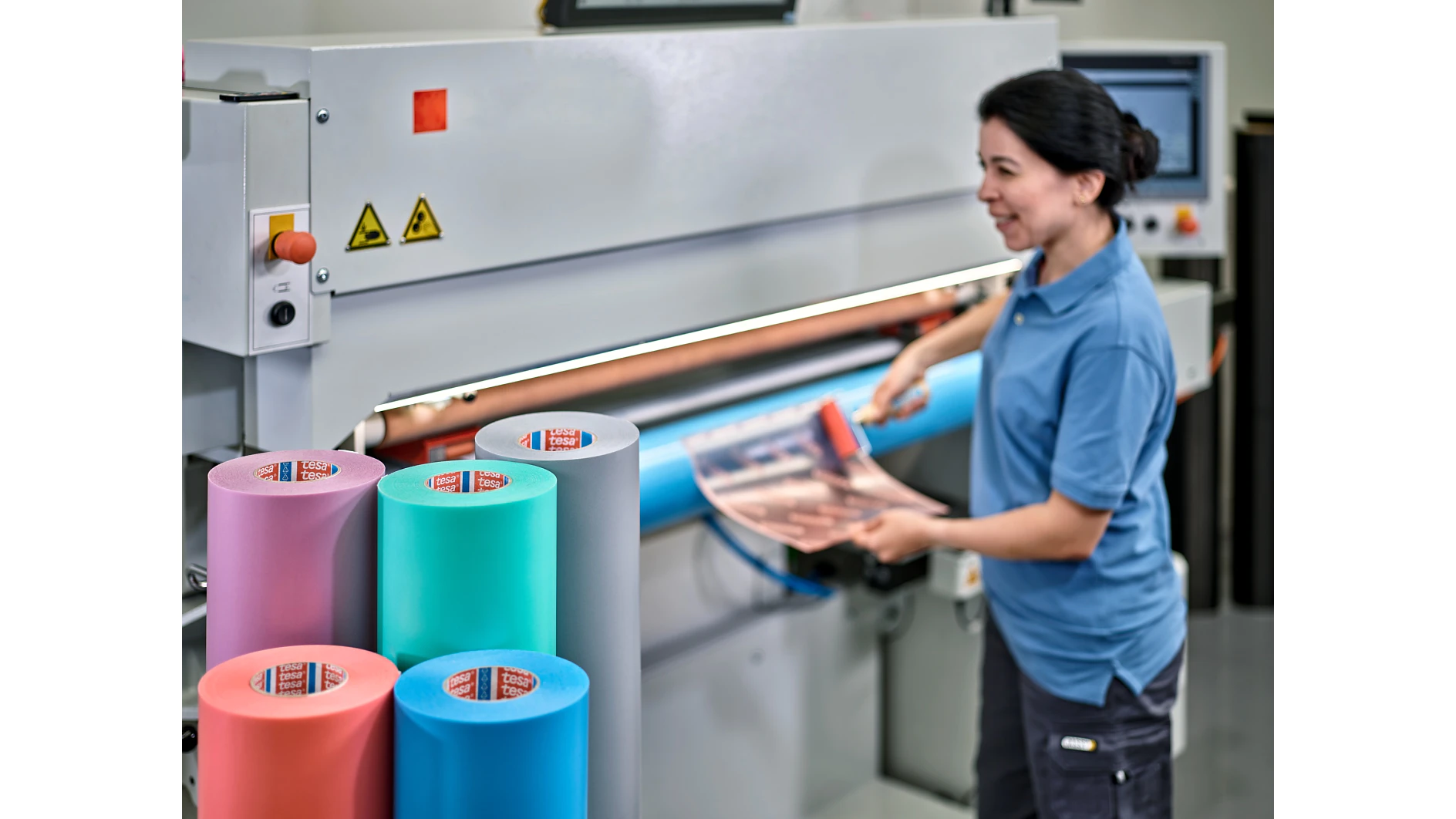 A person in a blue shirt operates a large industrial machine with multiple colored rolls of material, including green, pink, and blue. They are holding a roll of tesa tape against the machine. There are safety signs on the machine. (This text has been generated by AI)