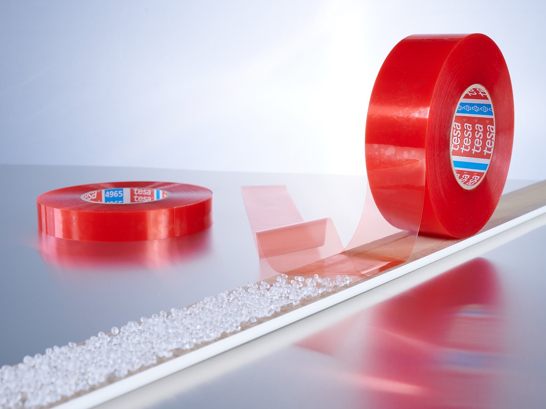 4965_7966_V1 Two rolls of red adhesive tesa tape are displayed against a light background. One roll is lying flat, and the other is standing upright with a strip of tesa tape partially unrolled and stretched out along a line of small white granules. (This text has been generated by AI)