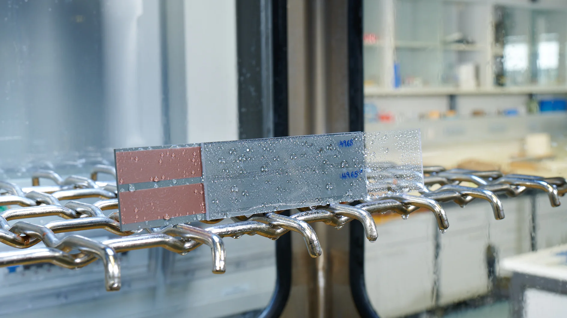 A metal strip with water droplets is placed on a rack inside a laboratory setting, secured with tesa tape. The strip has markings in blue text. The background showcases glass shelves and counters with laboratory equipment and containers. (This text has been generated by AI)