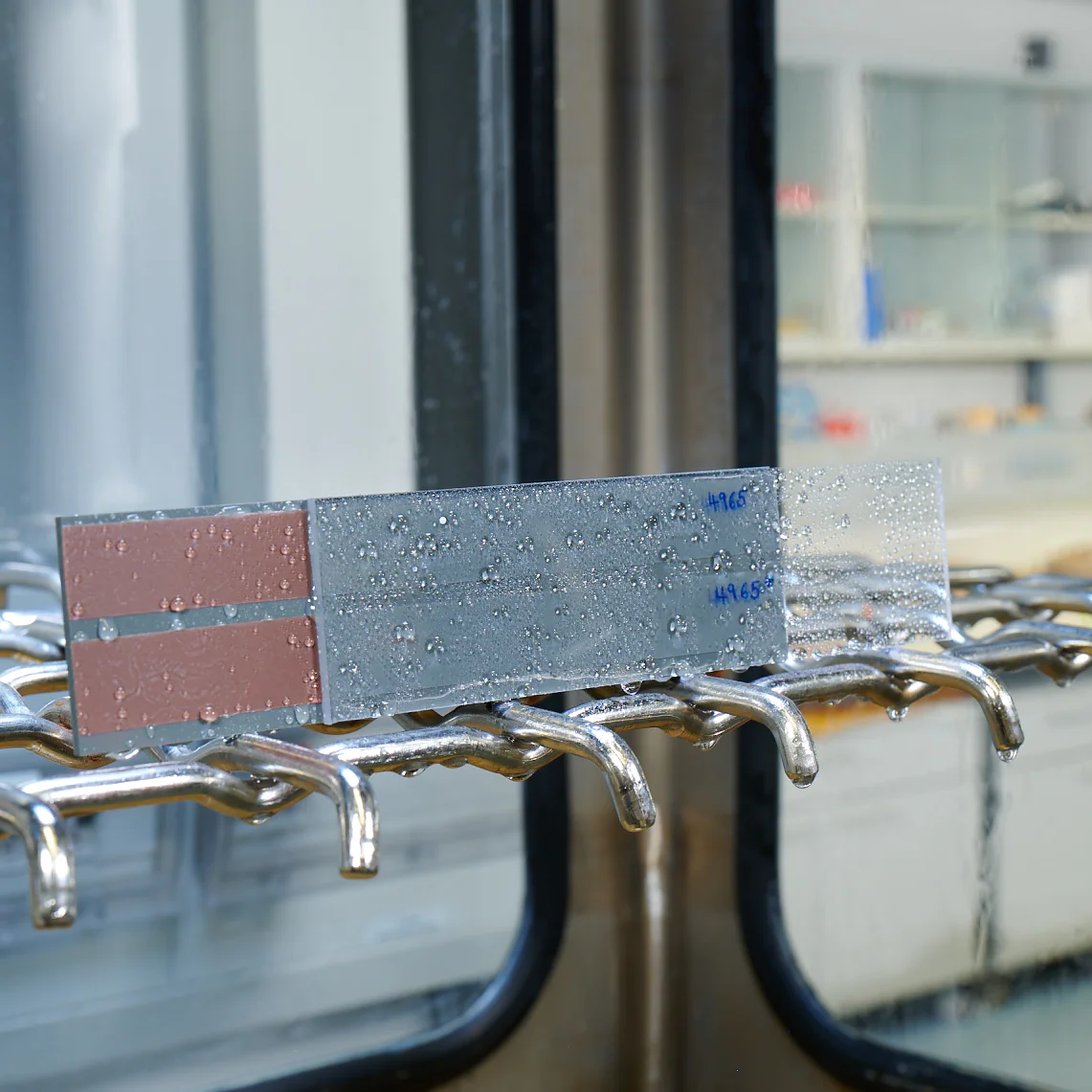 A metal strip with water droplets is placed on a rack inside a laboratory setting, secured with tesa tape. The strip has markings in blue text. The background showcases glass shelves and counters with laboratory equipment and containers. (This text has been generated by AI)