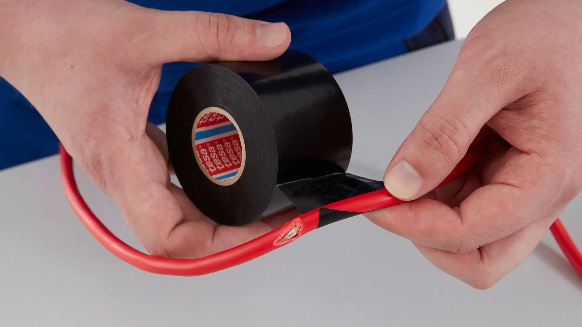 A person is holding a roll of black electrical tesa tape and wrapping it around a damaged red cable on a gray surface.