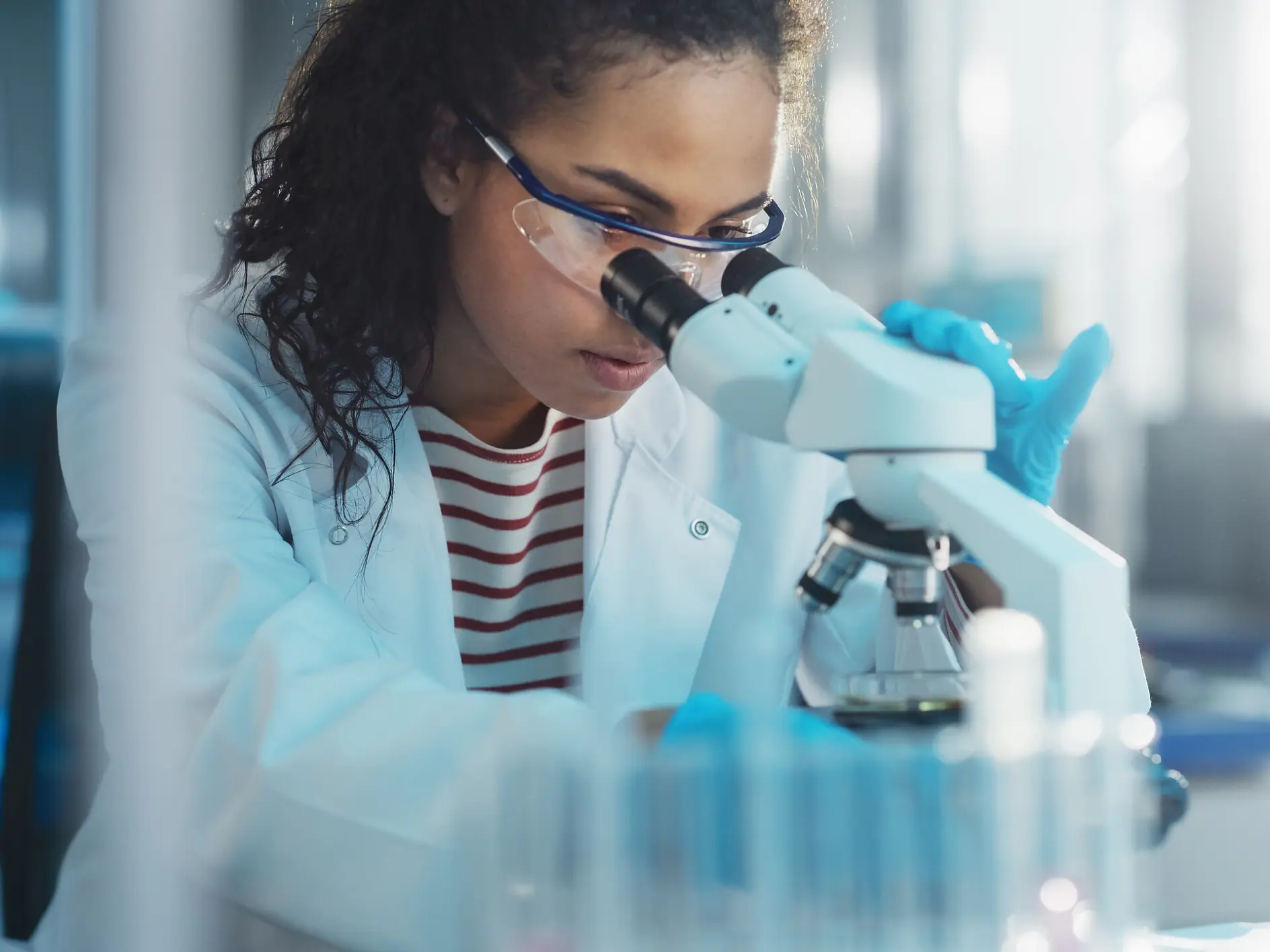 scientist wearing blue gloves using a microscope in a bright laboratory setting with test tubes nearby tesa (This text has been generated by AI)